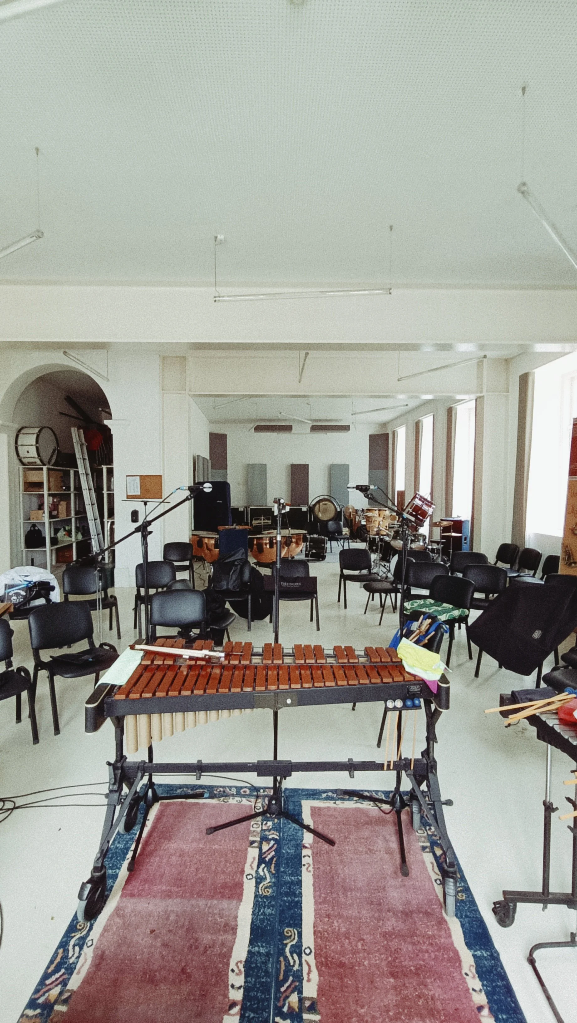 Music rehearsal room with chairs arranged in a semi-circle, music stands, a xylophone, and various percussion instruments set up at the front. The room has large windows on the right and soundproofing panels on the back wall.
