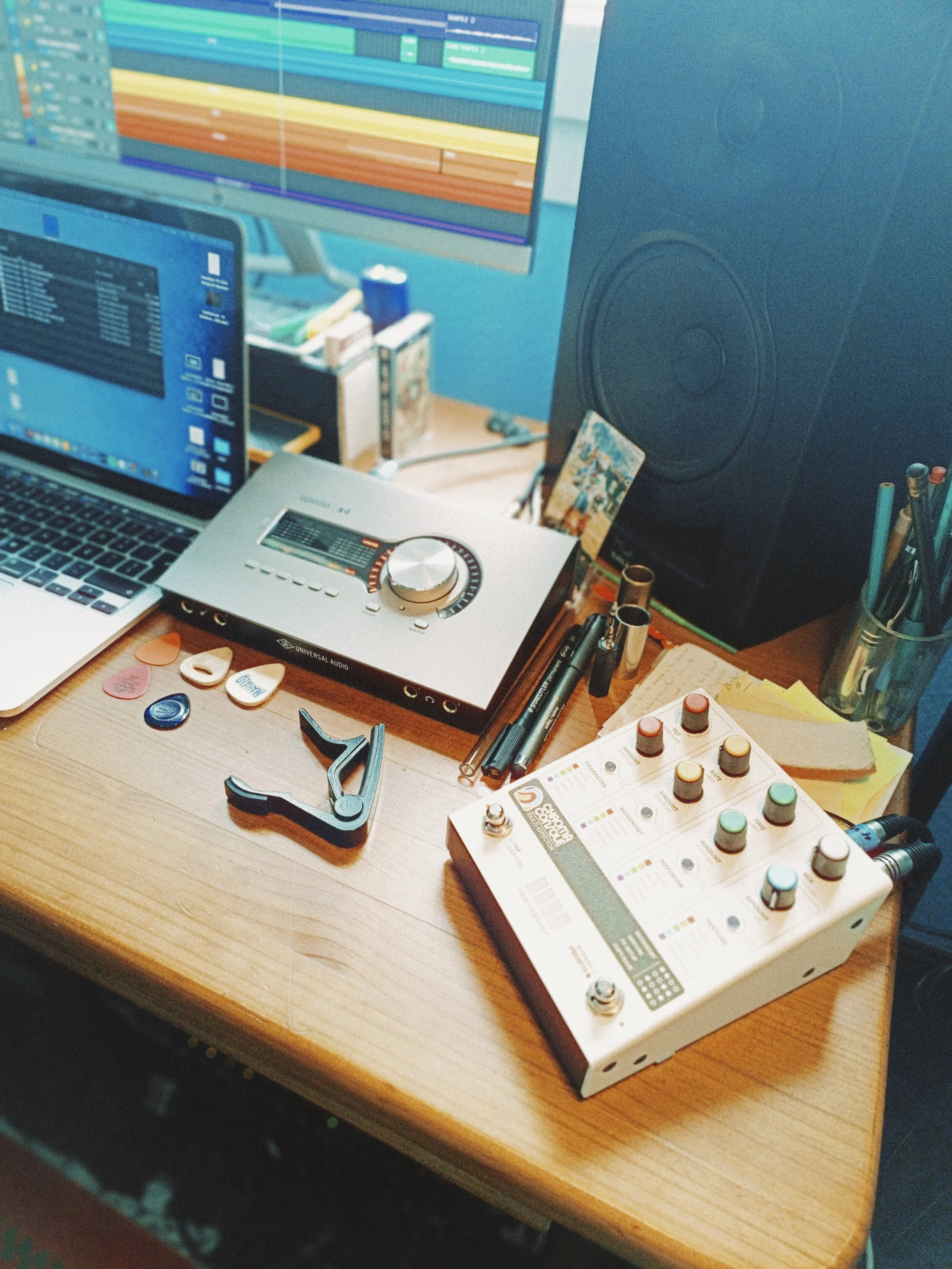 A desk with audio recording and editing equipment including a computer monitor with audio tracks, a laptop, a multimedia controller, a large speaker, and various small objects like guitar picks, pens, and a small notebook.