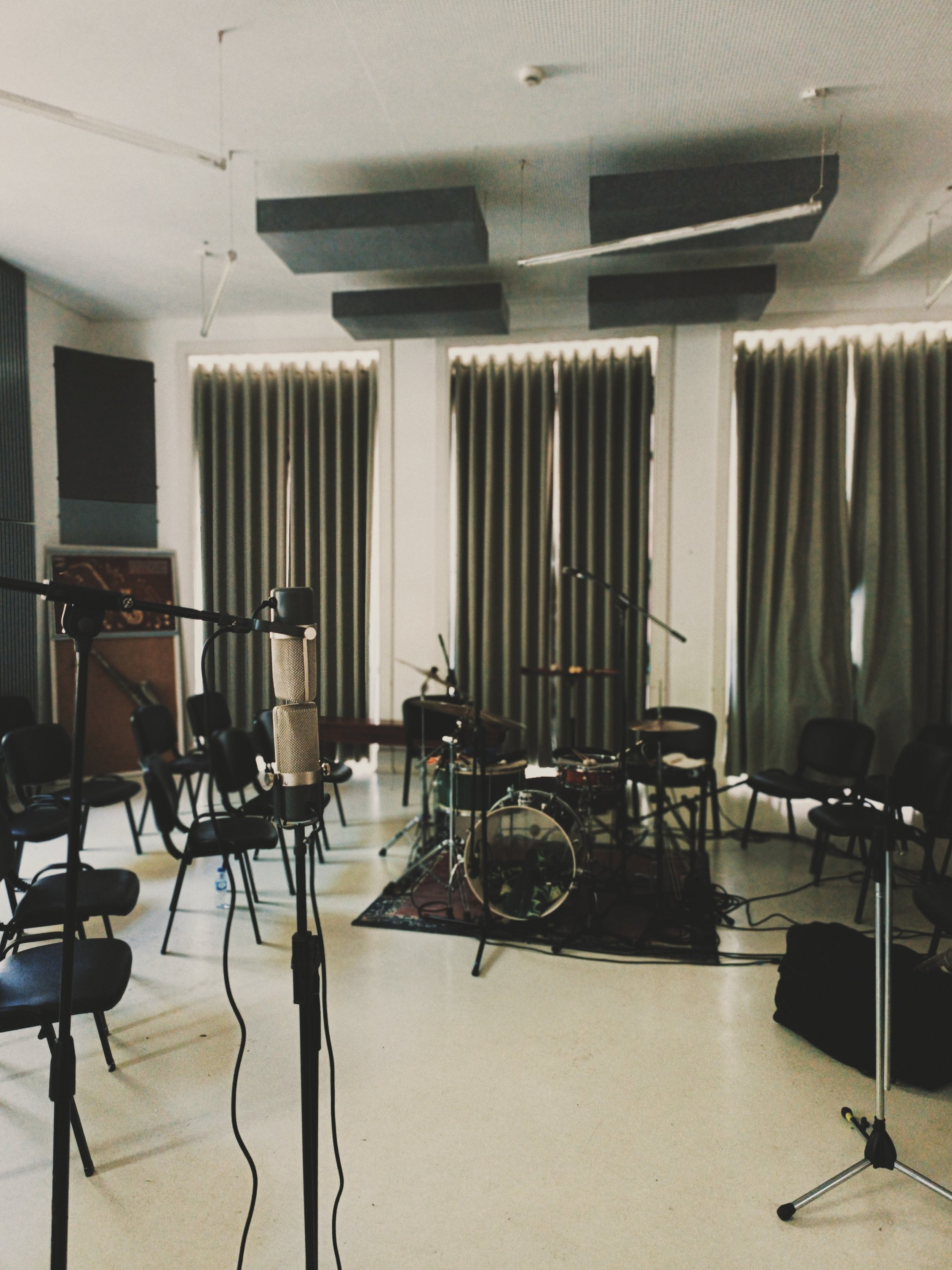 Empty room set up for music recording or rehearsal with multiple microphones on stands, a drum set on a rug, chairs, and soundproofing panels on the ceiling, with long curtains along windows.