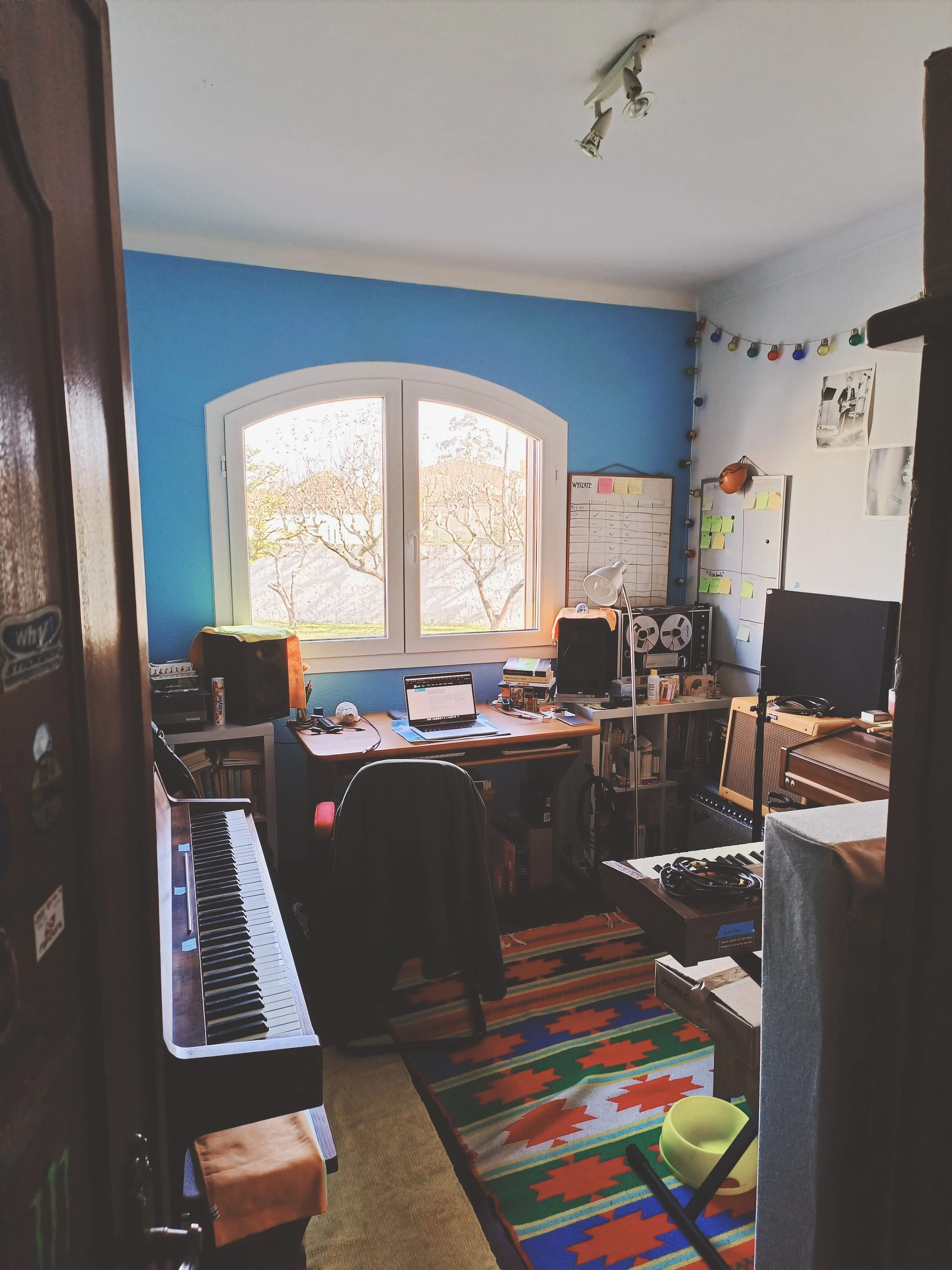 A cozy home office with a large arched window, blue accent wall, and a window sill with sunlight coming through. The room contains a keyboard on a desk, a laptop, speakers, and various office supplies. There are colorful string lights in the corner, 