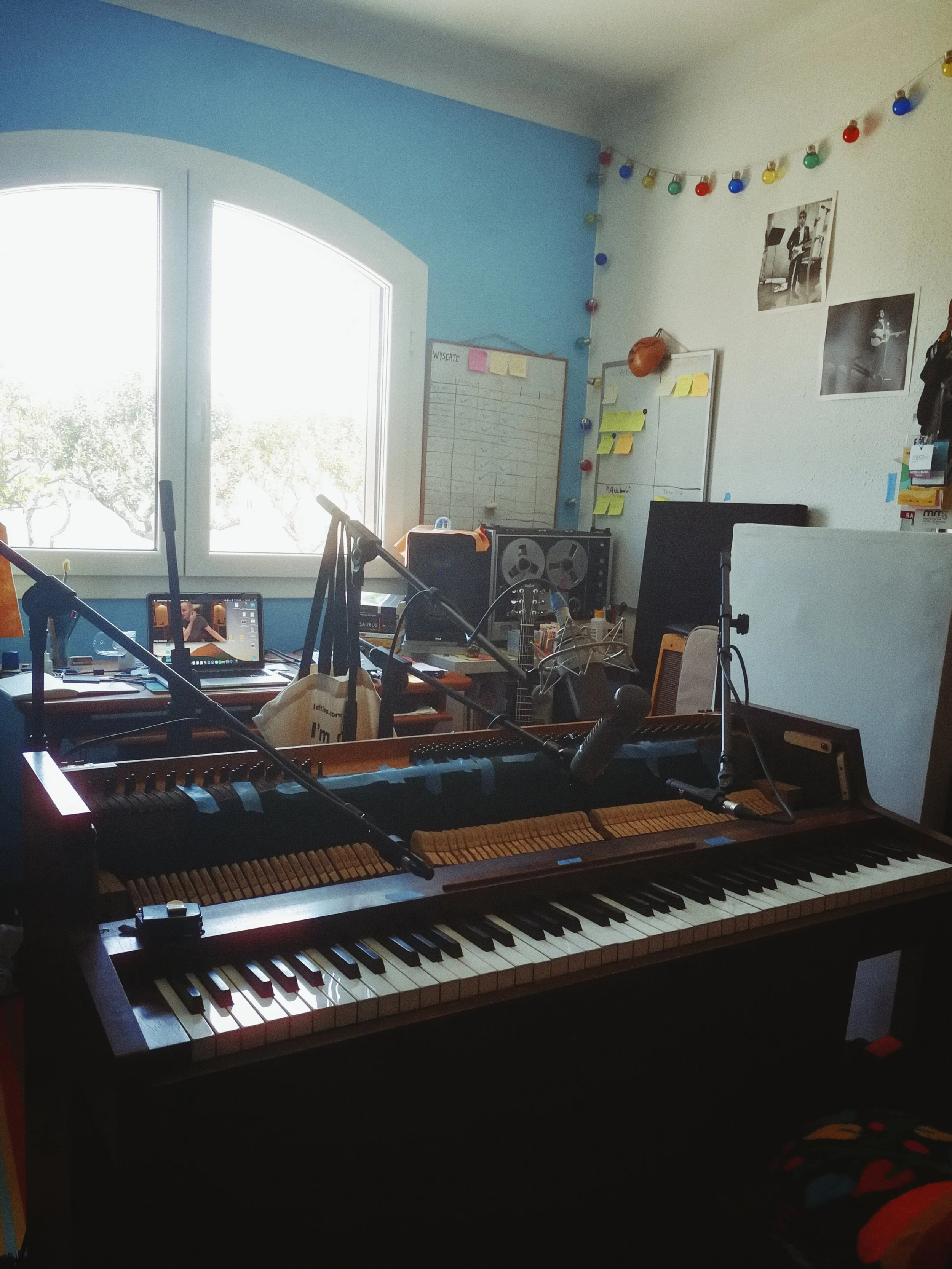 An indoor room with a large window on the left, a keyboard piano in the foreground, and various electronic equipment, posters, and decorations on the walls. The room has a blue and white color scheme with string lights hanging across the white wall.