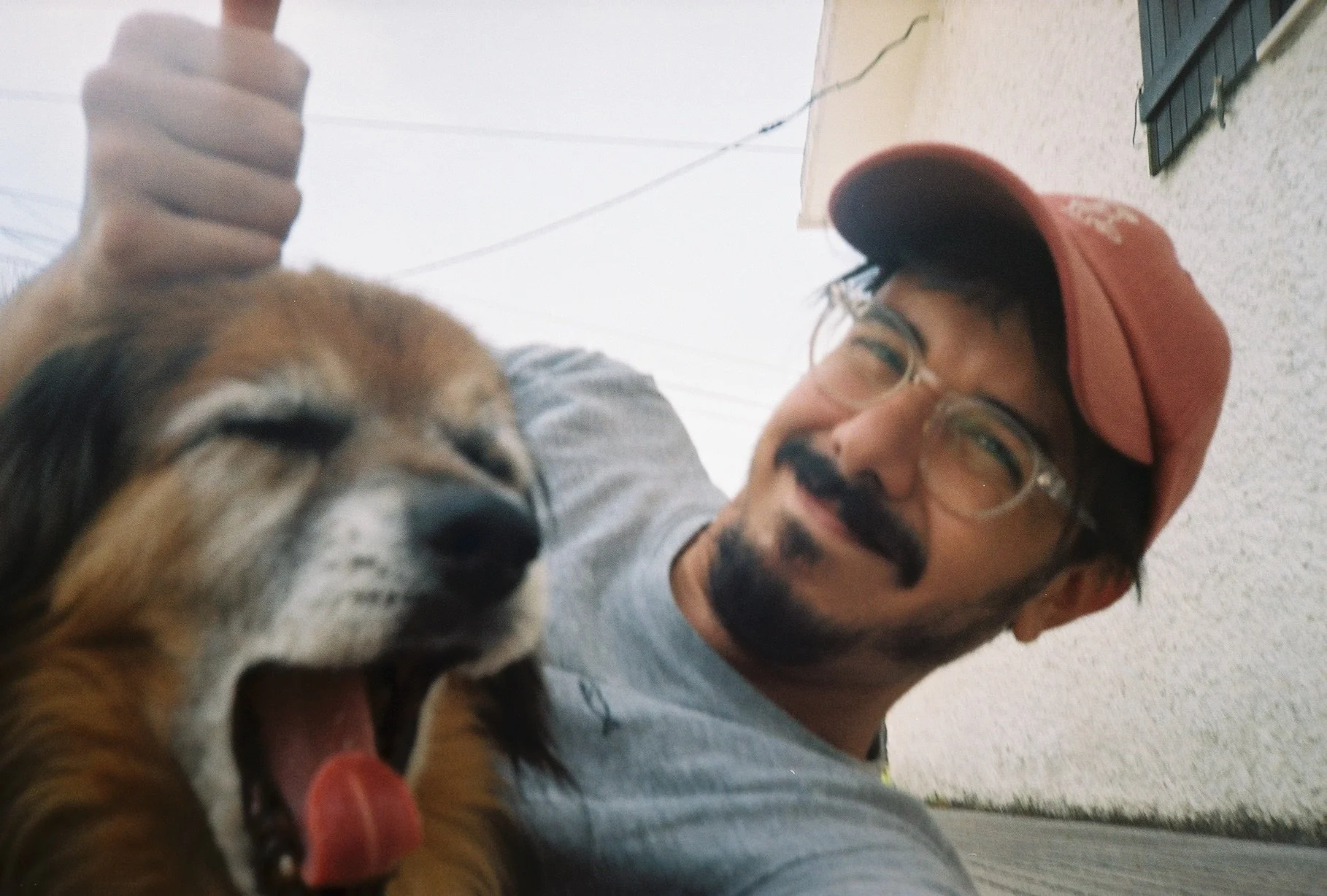 A man with glasses and a beard wearing a red cap is smiling and holding a large barking dog, both looking at the camera.