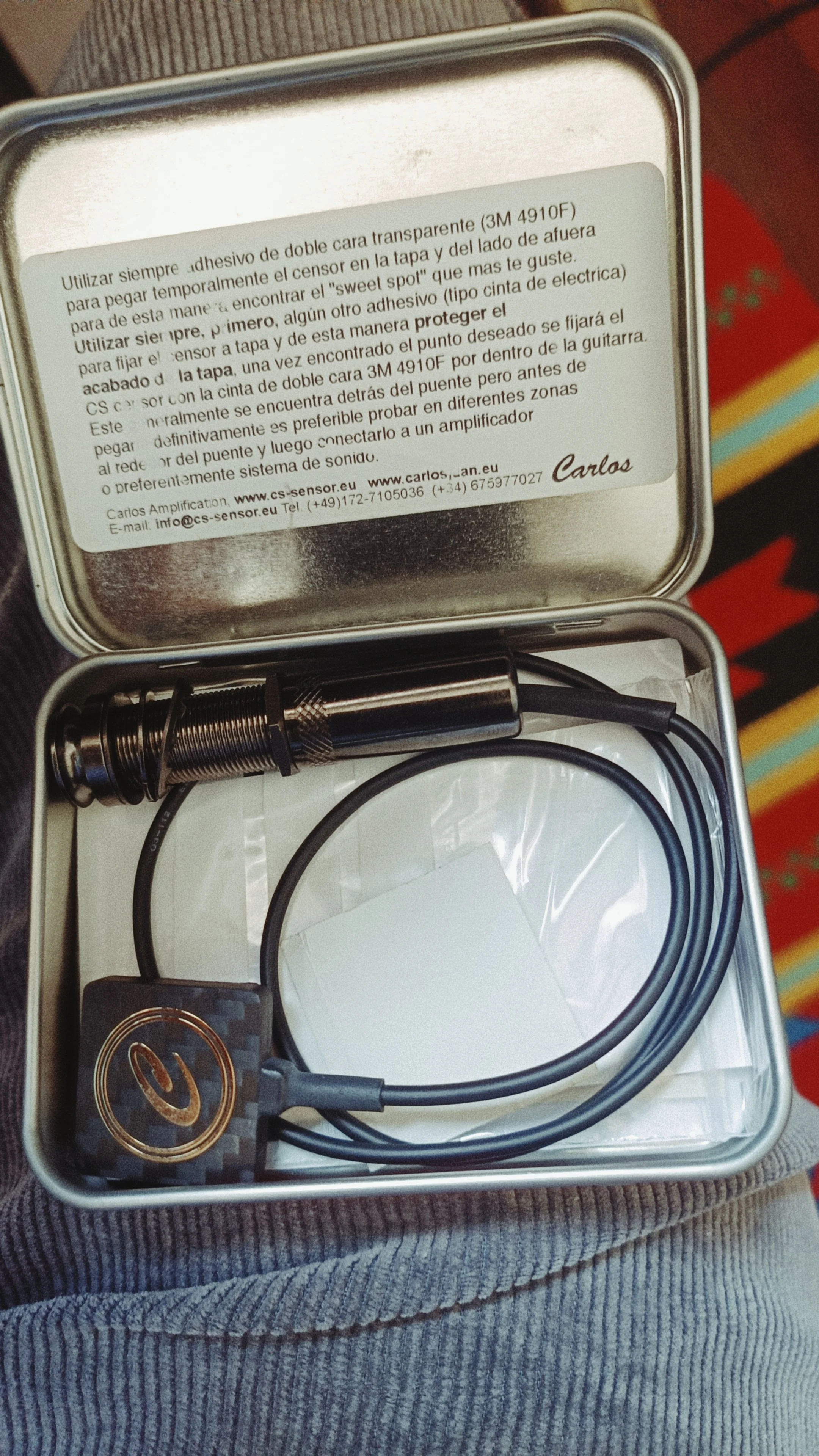 Open metal box containing a coil spring, a black cable, and a circuit board with a power button symbol, on a person's lap with gray striped pants.