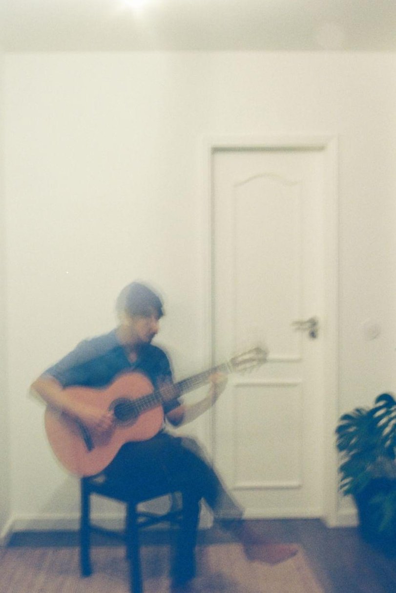A person sitting on a chair playing an acoustic guitar in a room with plain white walls and a closed door.