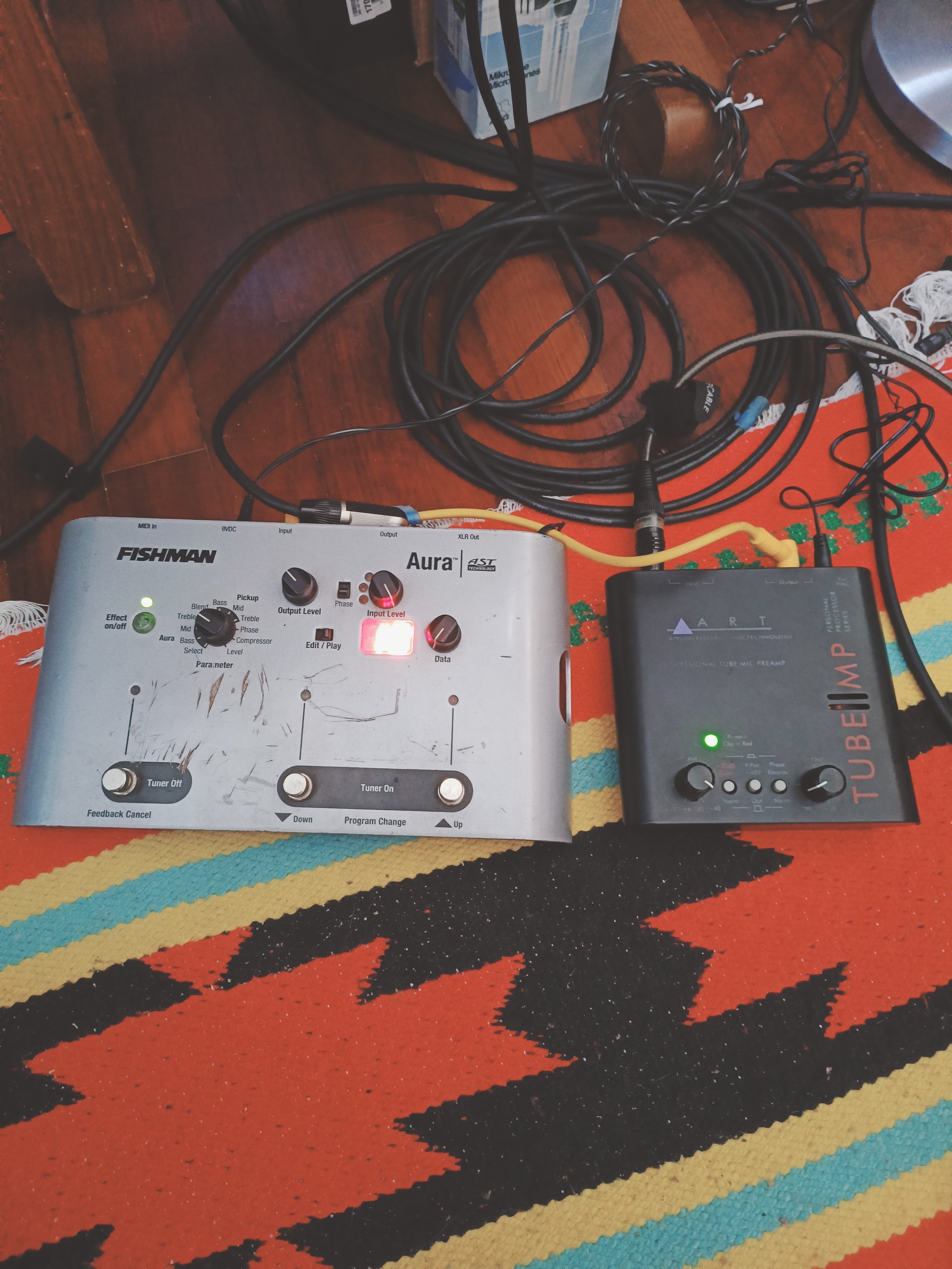A digital audio interface device, possibly a Fishman Aura, with various knobs and controls, connected to other audio equipment with multiple black cables. The equipment is placed on a colorful patterned rug on a wooden floor.