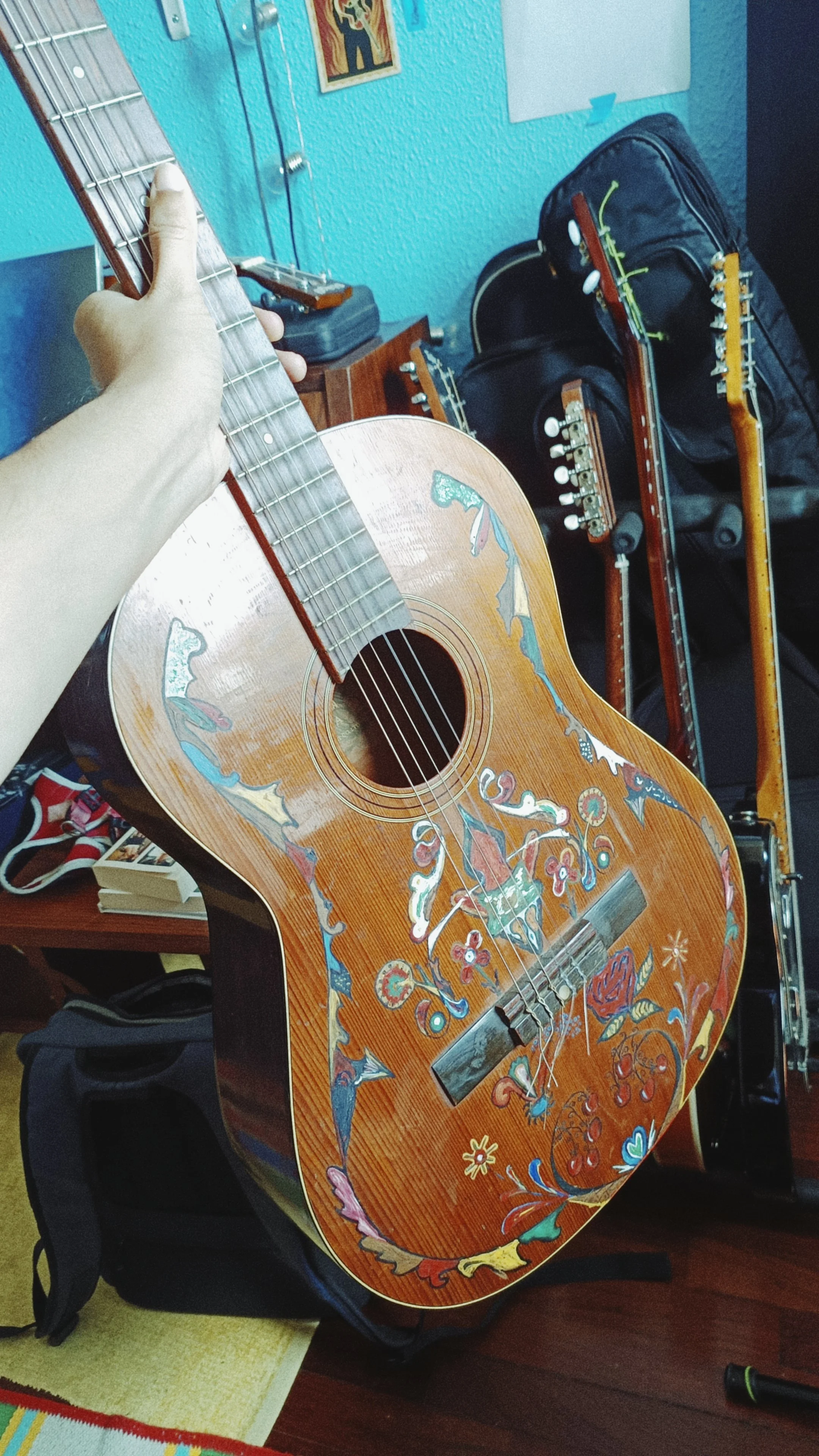 Decorated acoustic guitar resting on a stand, with a colorful painted design on its body. In the background, two more guitars, a guitar case, and a wall with artworks.