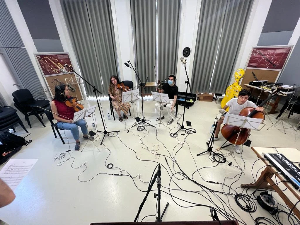 A group of five musicians performing in a recording studio with various instruments and microphones, with cables on the floor.