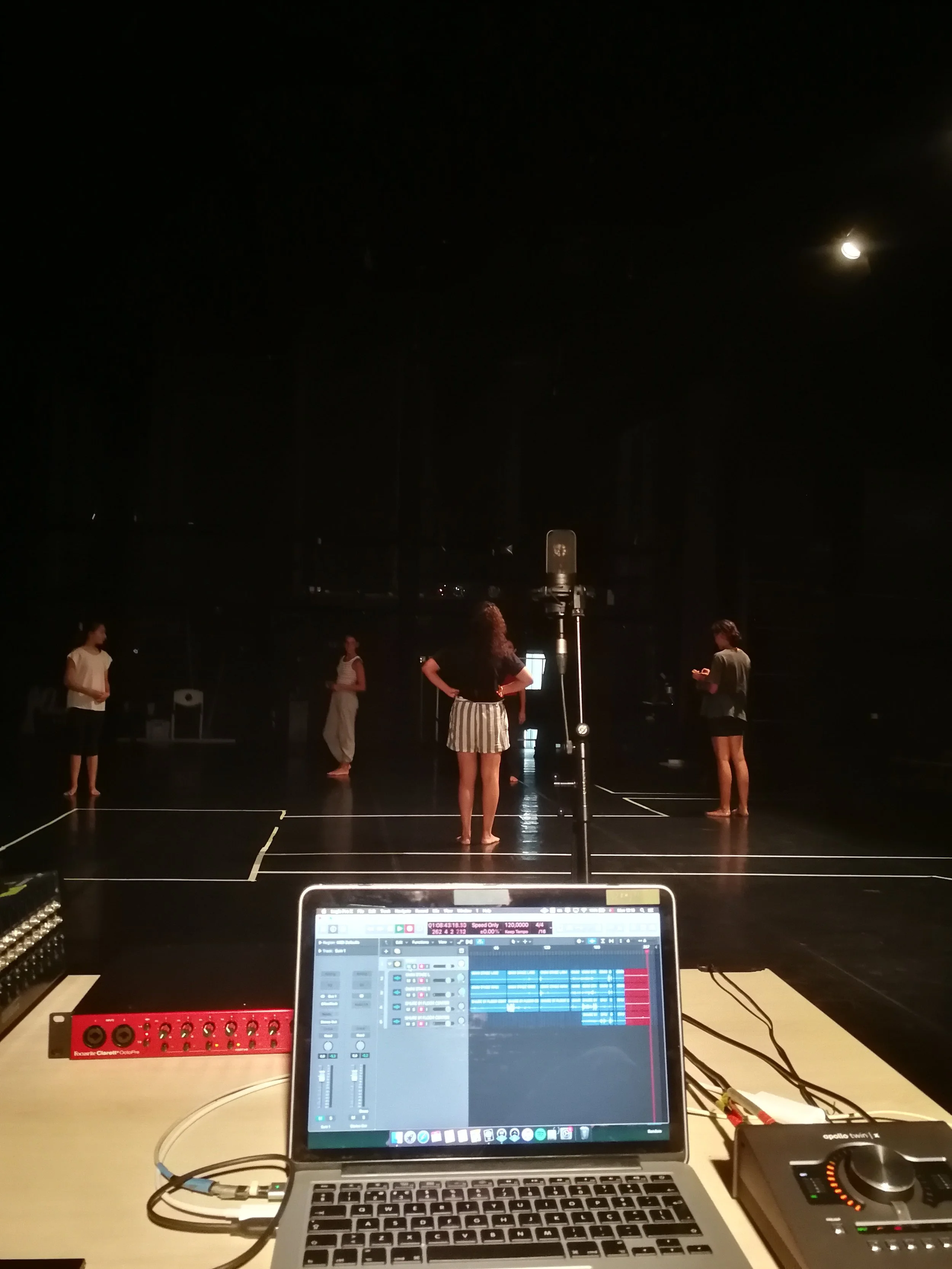 A photo showing a laptop with music production software open, surrounded by audio equipment, in front of a stage with dancers practicing under a dark sky with the moon visible, during a night rehearsal.
