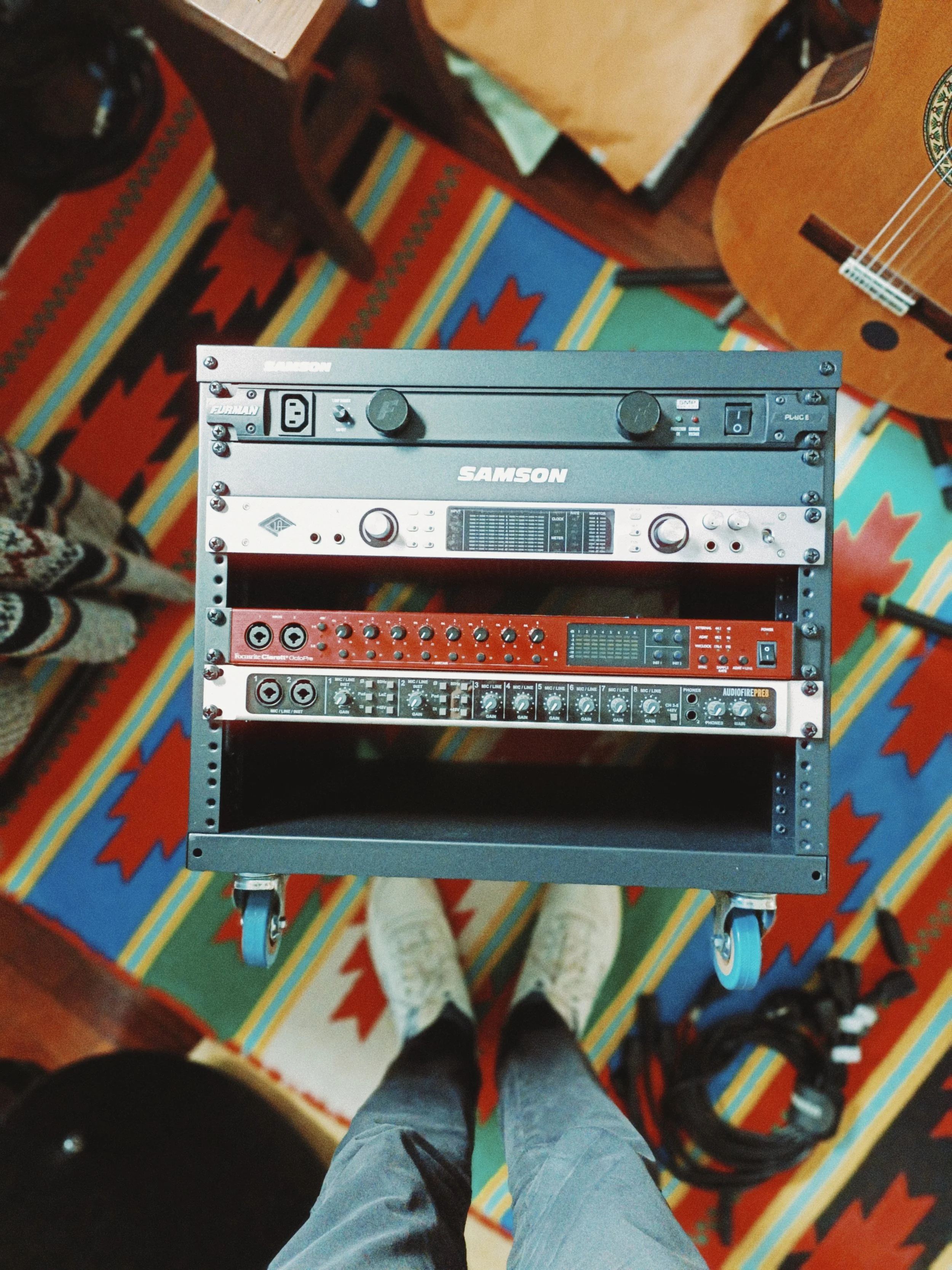 A person standing on a colorful southwestern patterned rug, wearing gray pants and sneakers, viewed from above, with a music equipment cart holding various audio devices.