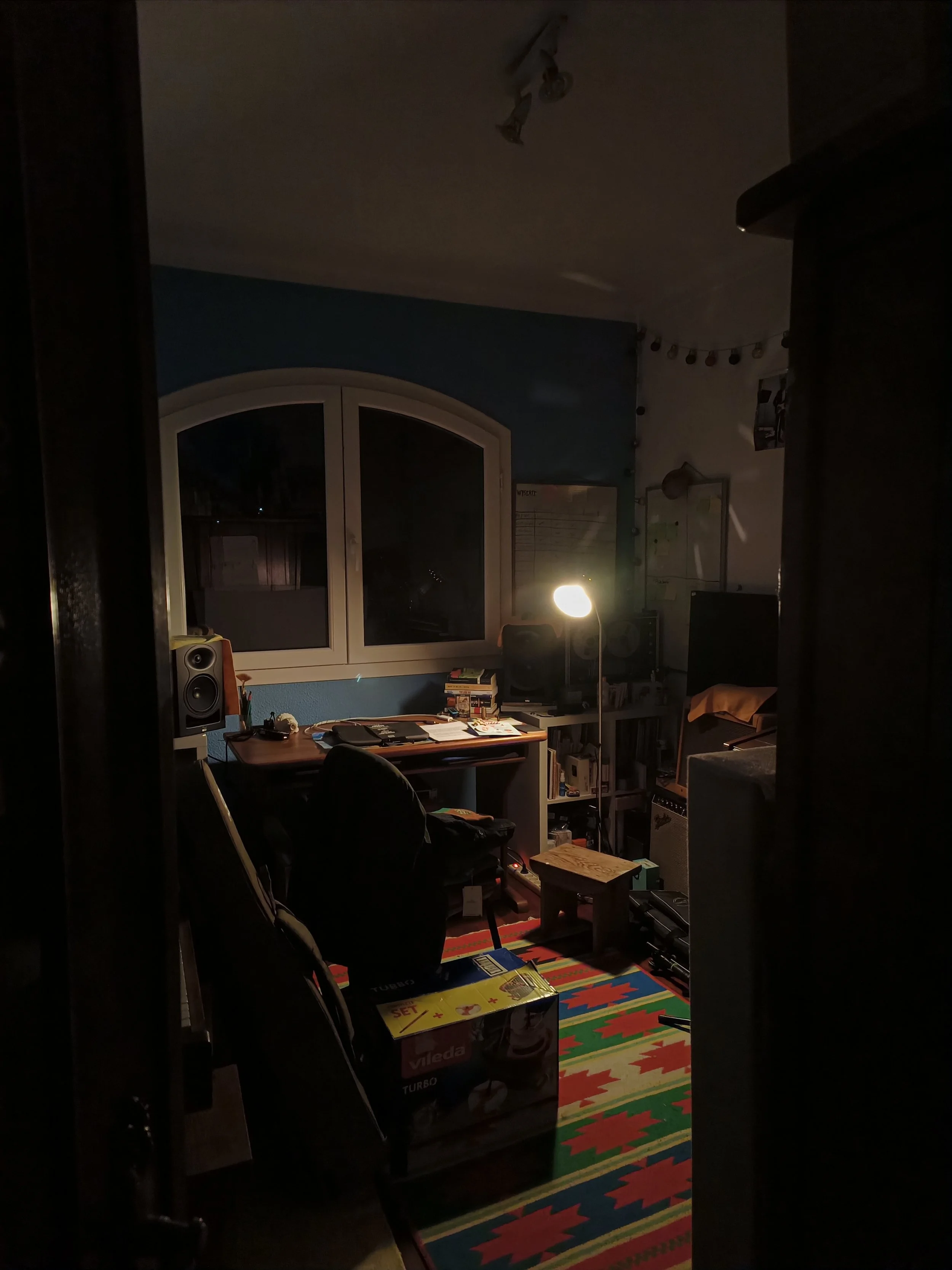 A dimly lit room with a window showing night outside, a desk with books and desk accessories, a speaker, a lamp, a colorful rug, and some furniture and boxes inside.