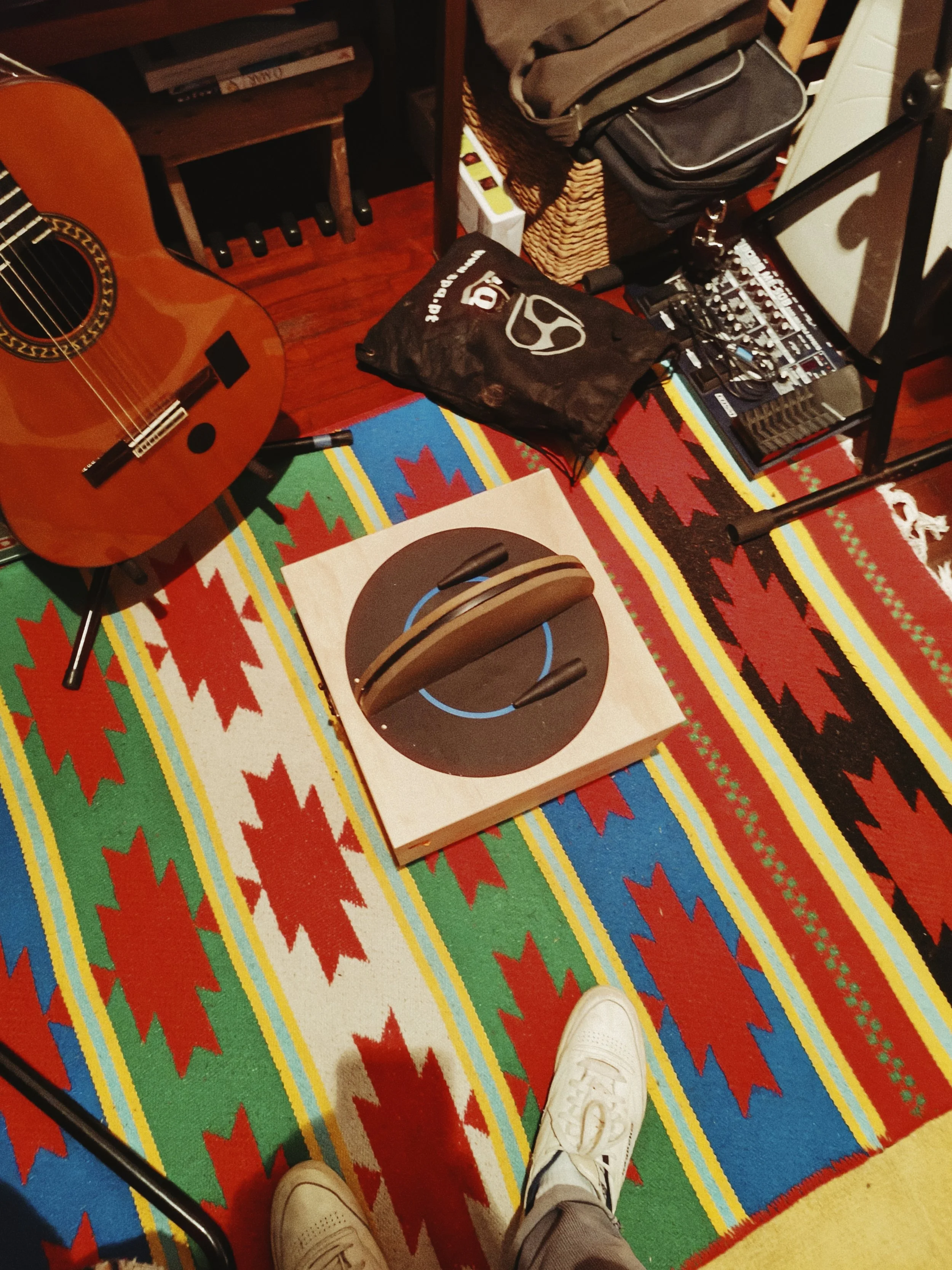 A colorful patterned rug with a red, green, blue, yellow, and white design. On the rug, there is a cardboard box and a pair of white sneakers. Part of an acoustic guitar is visible on the left side of the image. The person's feet are in the foregroun
