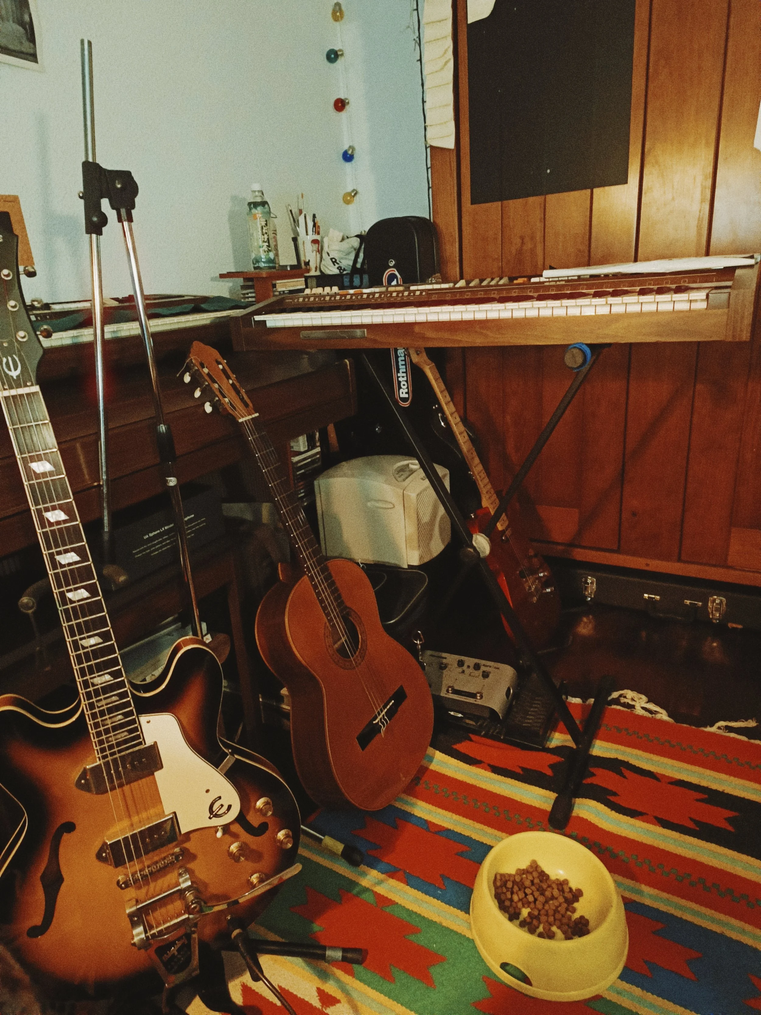 An indoor room with musical instruments including two guitars, a keyboard, and a music stand. There is a colorful striped rug on the floor, and a bowl of cereal on a yellow dish. The wall has a wood-paneled section and a light-colored section with so