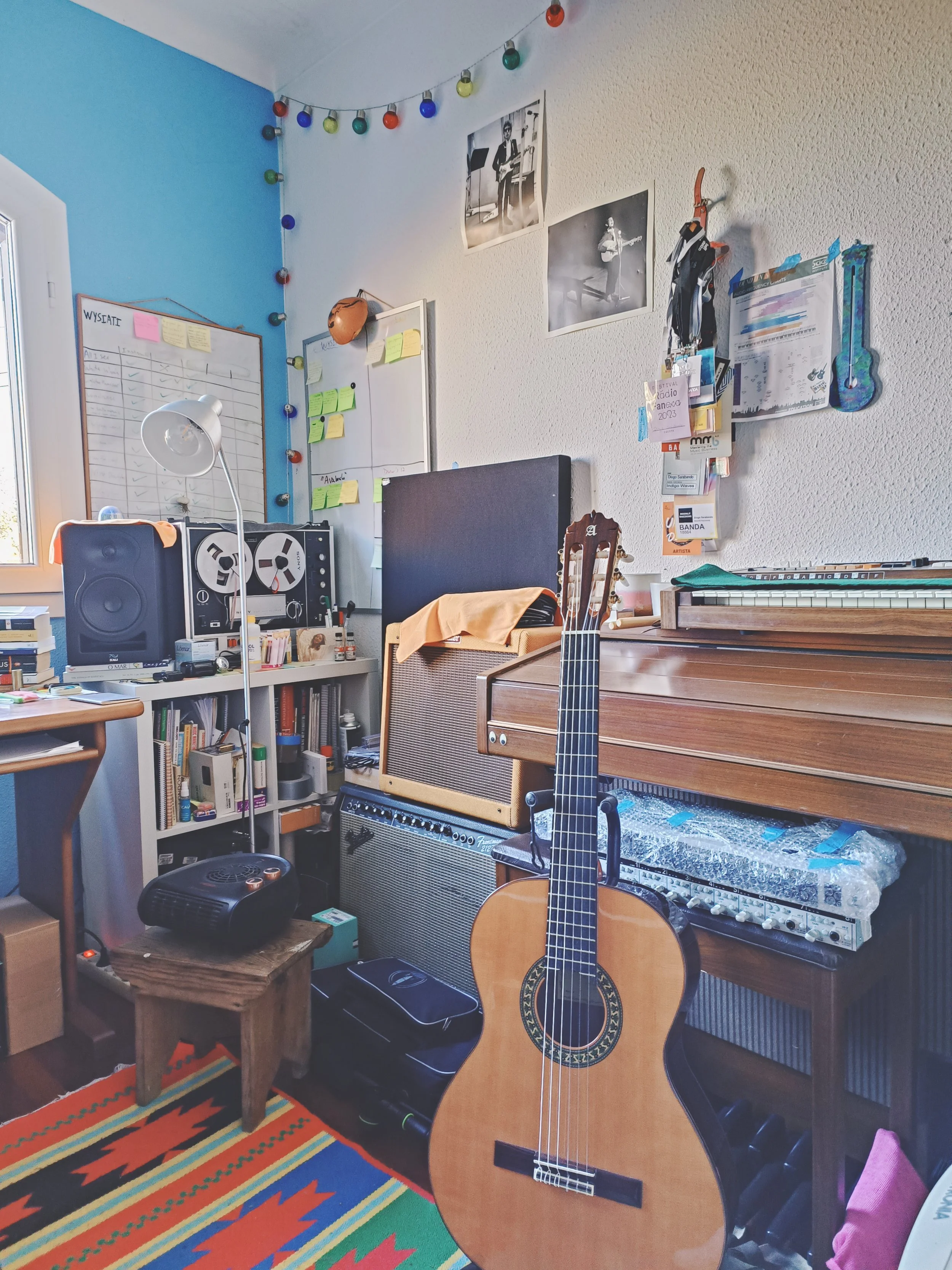 Room with musical instruments and decor: guitar, amplifier, shelves, posters, colorful string lights, and a patterned rug.
