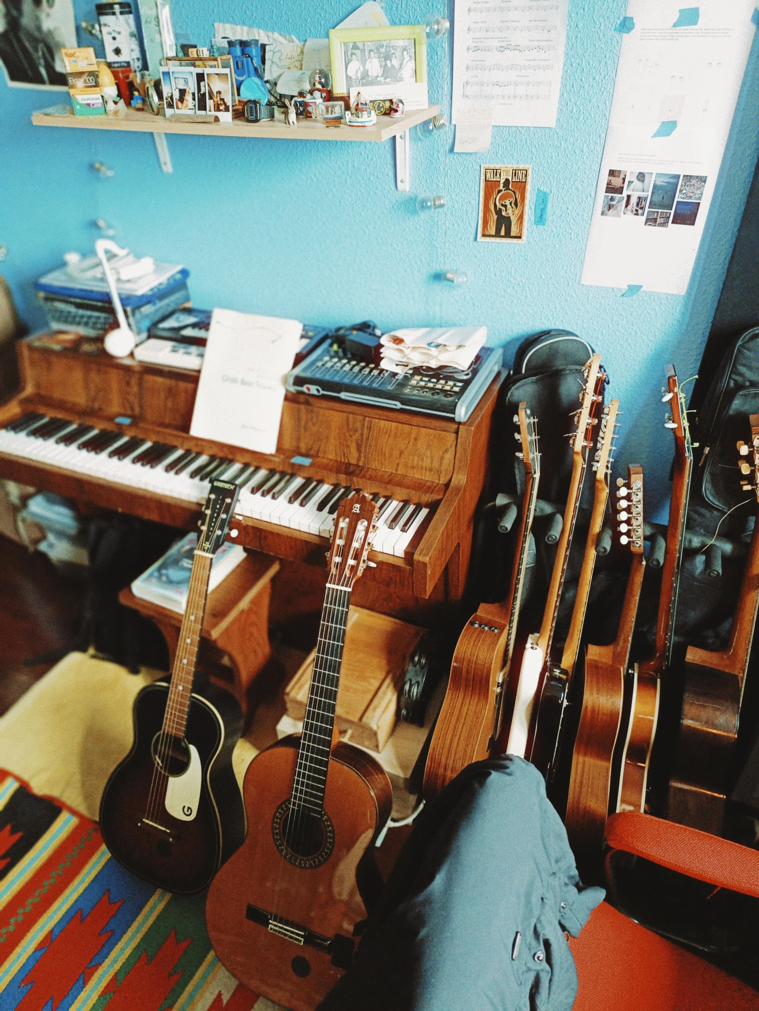 A room with musical instruments including a piano and several guitars, a desk with audio equipment, and a wall decorated with photographs, posters, and notes.