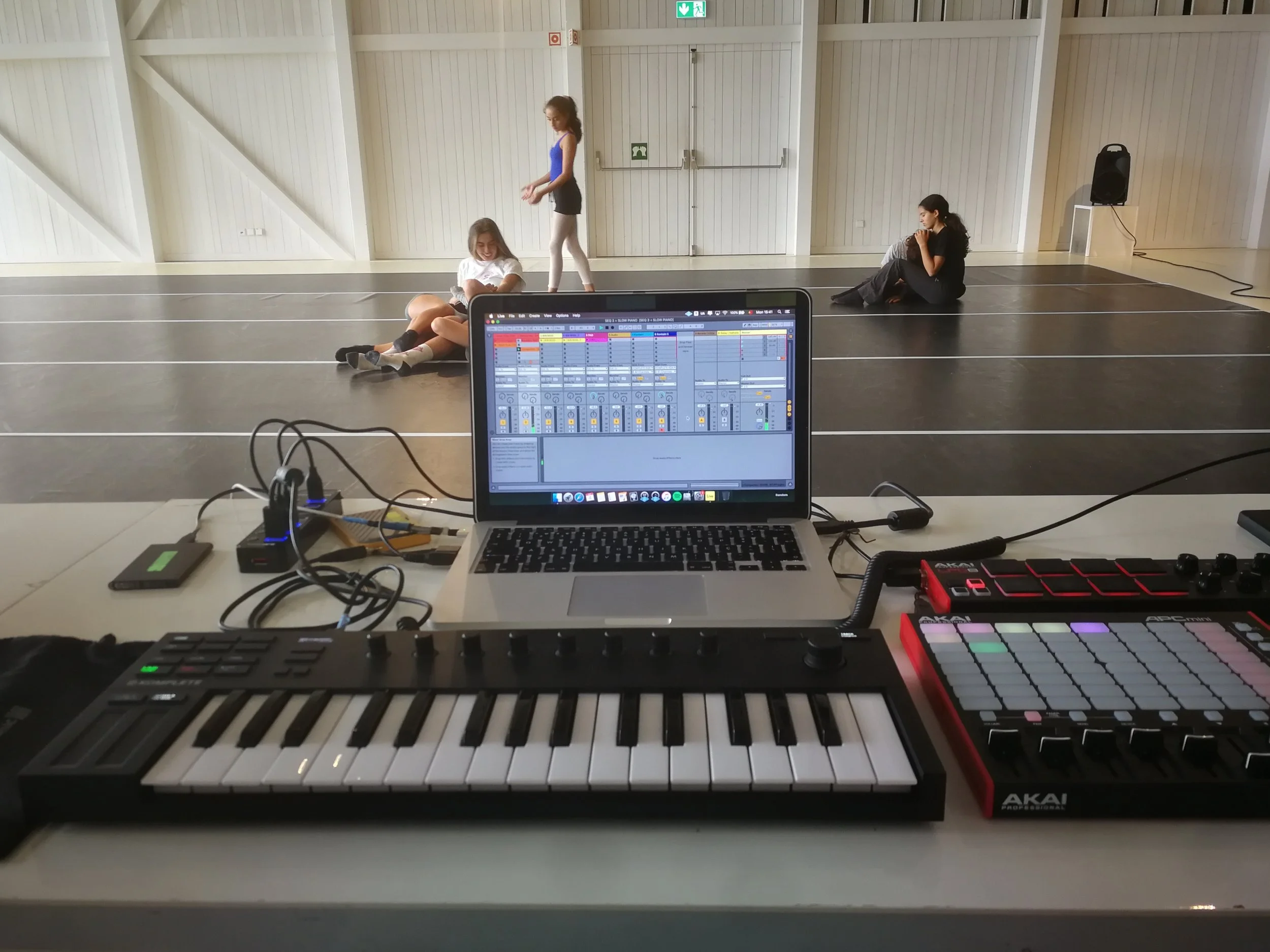 A spacious room with wooden walls, three girls sitting or standing on the floor, and musical equipment including a keyboard, a laptop with music editing software, and electronic music controllers.