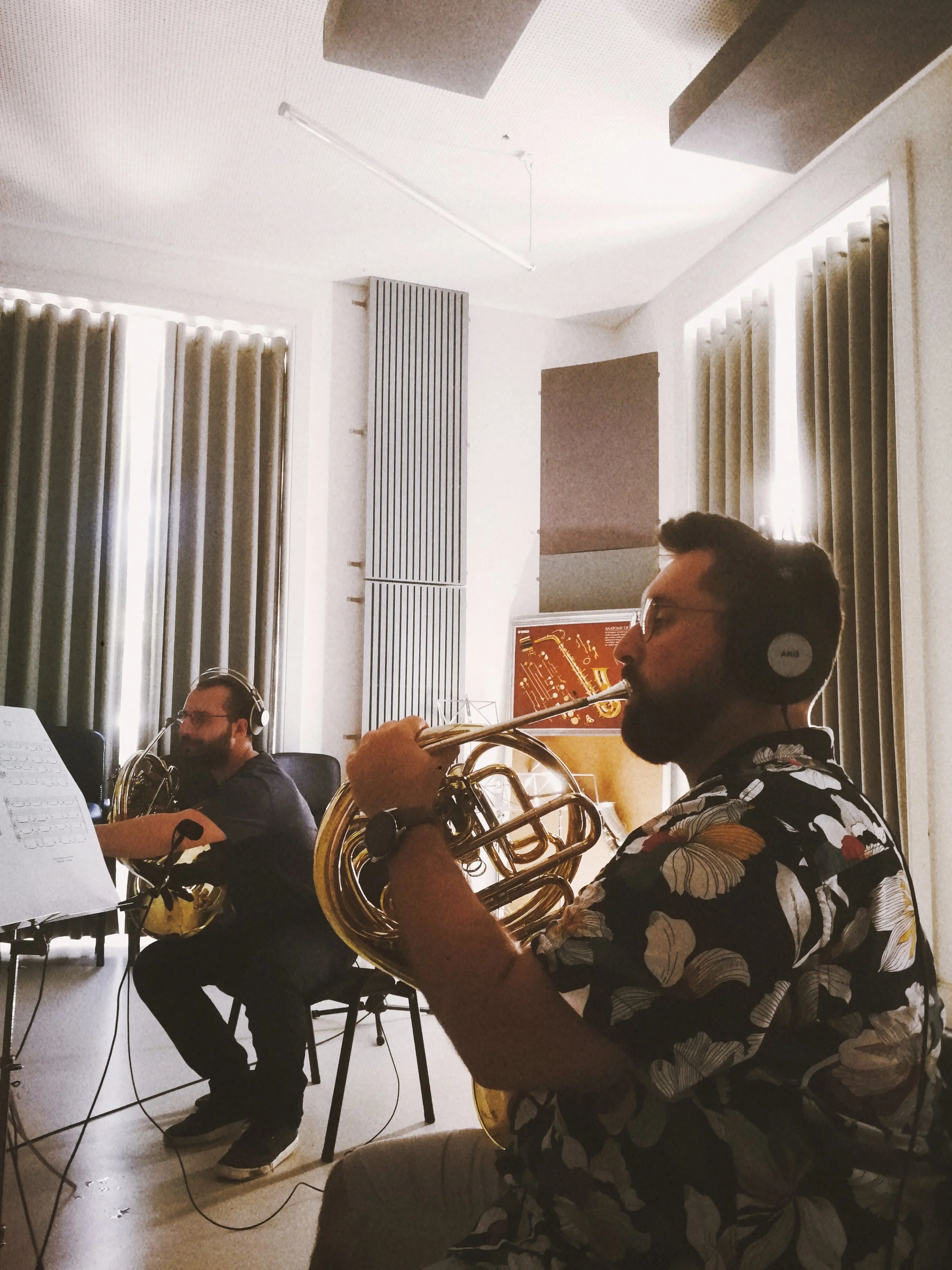 Two musicians playing French horns in a music studio, with sheet music, computers, and soundproof windows in the background.