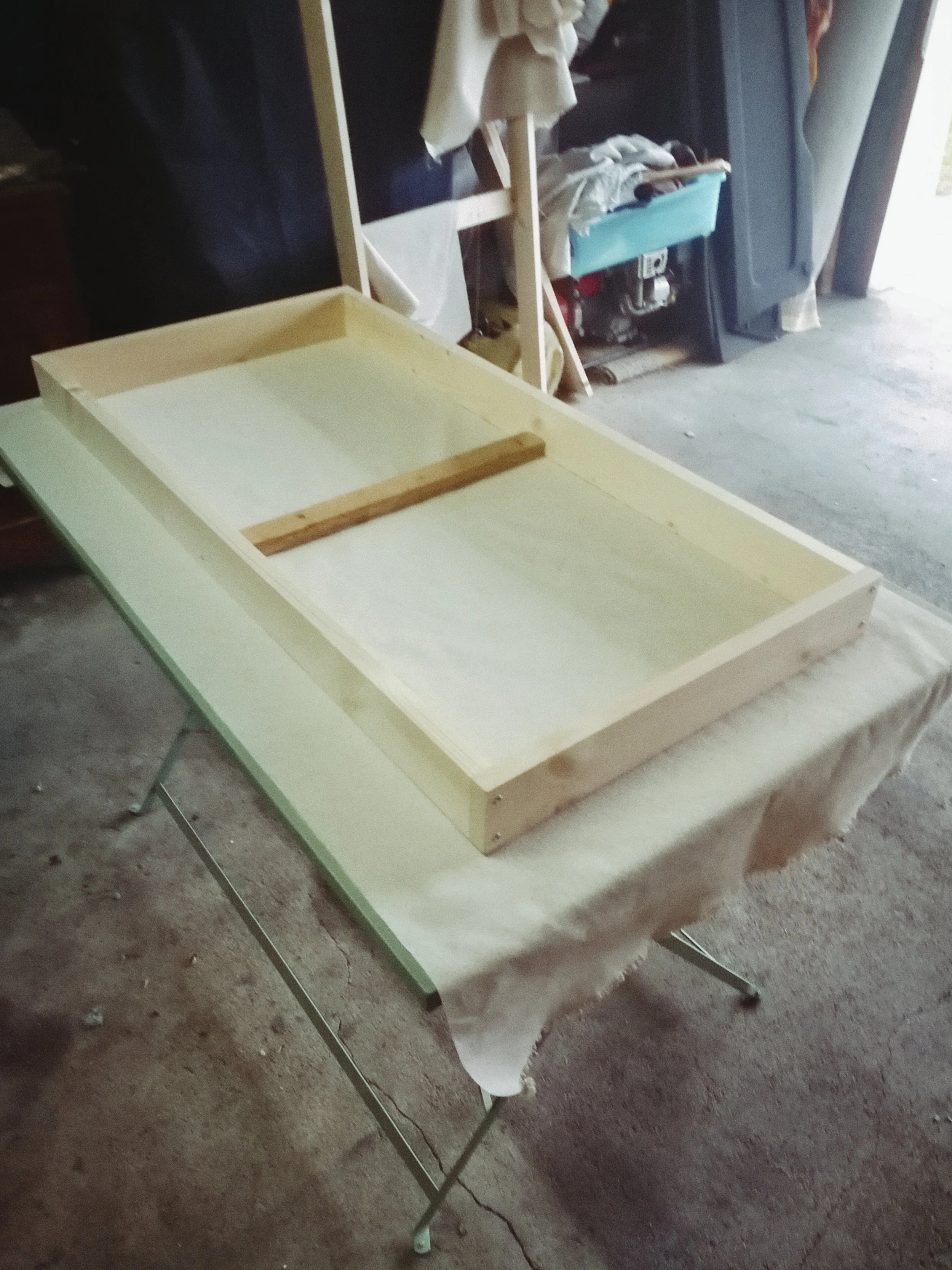 Unfinished wooden frame on a table, with a workspace and various tools in the background, including a blue storage container and a black cabinet.