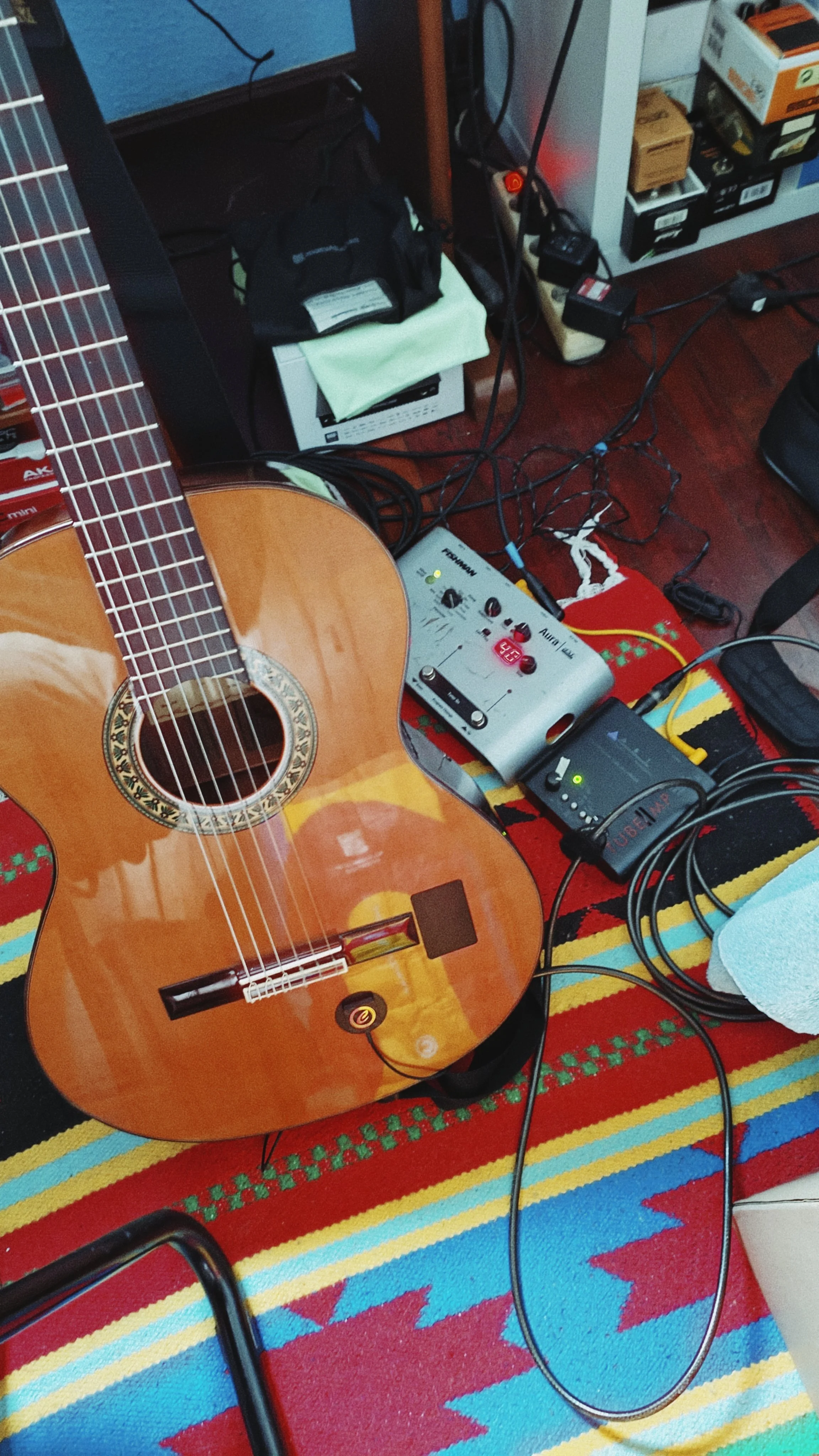 An acoustic guitar on a colorful striped rug, surrounded by electronic music equipment, wires, and a power strip.