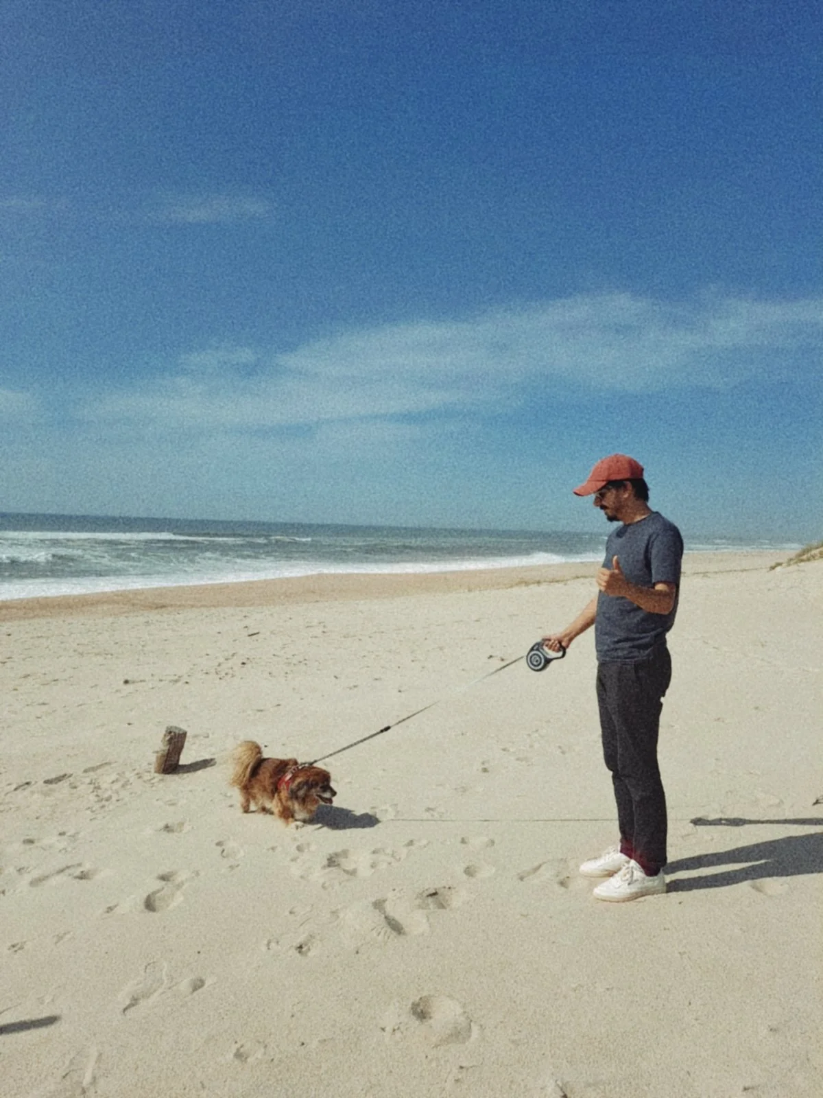 A man walking his small dog on a sandy beach. The man is wearing a pink baseball cap, sunglasses, a blue T-shirt, black pants, and white sneakers. The dog appears to be a Pomeranian breed and is on a leash. The beach is wide with footprints in the sa