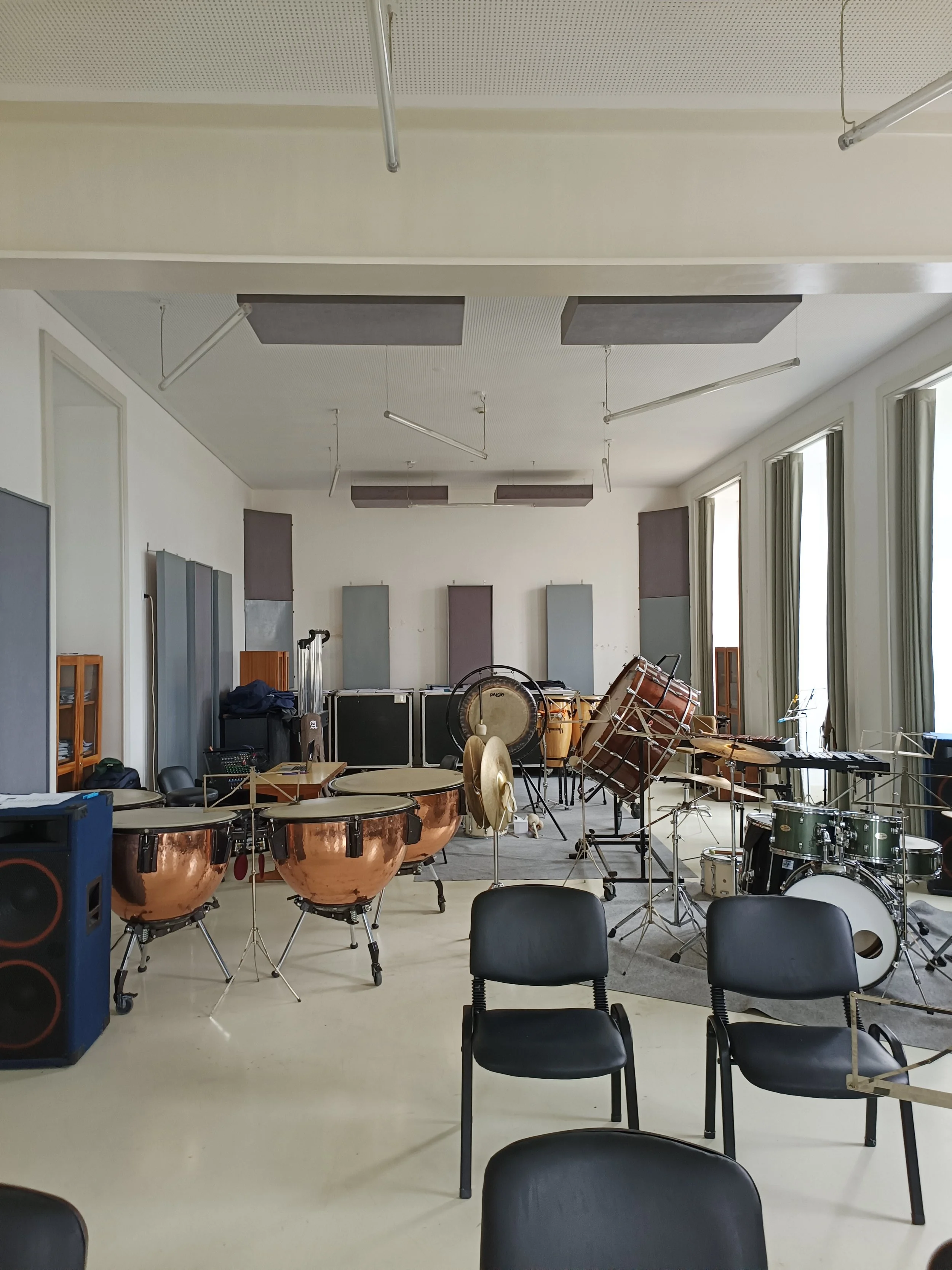 Music rehearsal room with various percussion instruments including chimes, drums, cymbals, and a xylophone set, along with chairs and audio equipment, illuminated by natural light from tall windows.