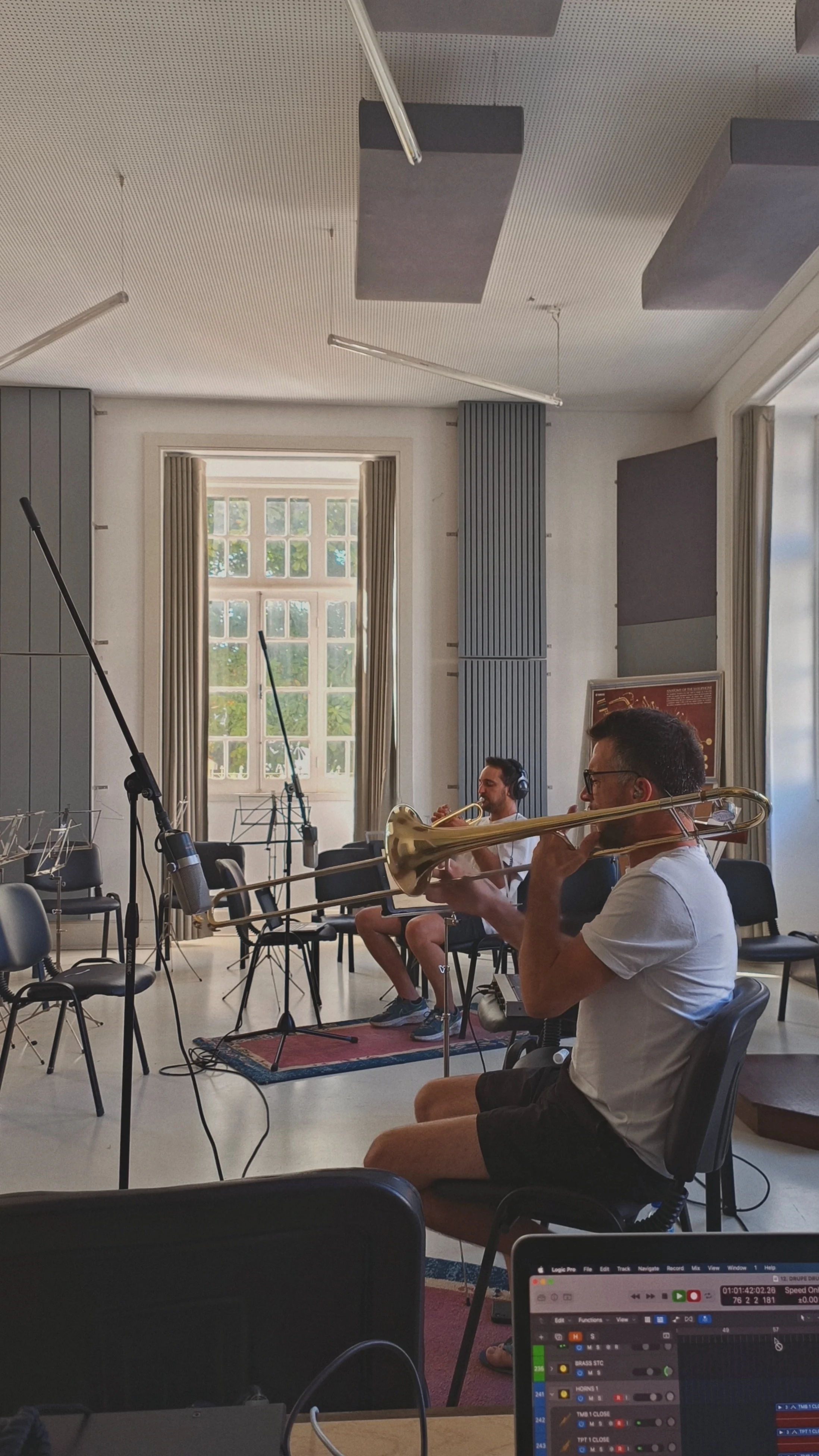 Two musicians in a recording studio playing trumpet and trombone, with music stands and chairs around, a large window in the background, and a laptop showing audio editing software in the foreground.