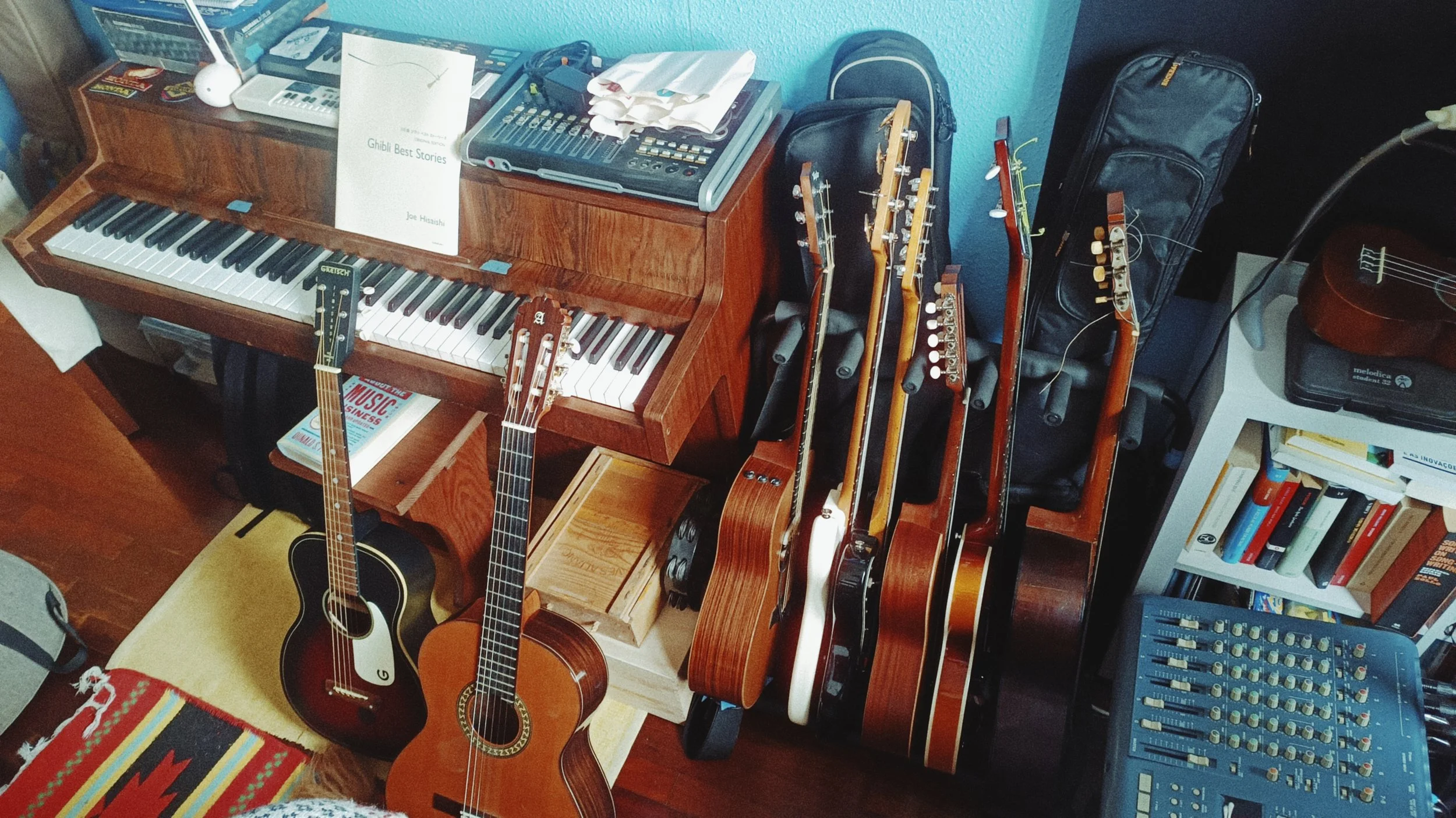 A room with musical instruments including two acoustic guitars, a wood piano with sheet music, and several electric guitars, along with a music book, bookshelves, and musical equipment.