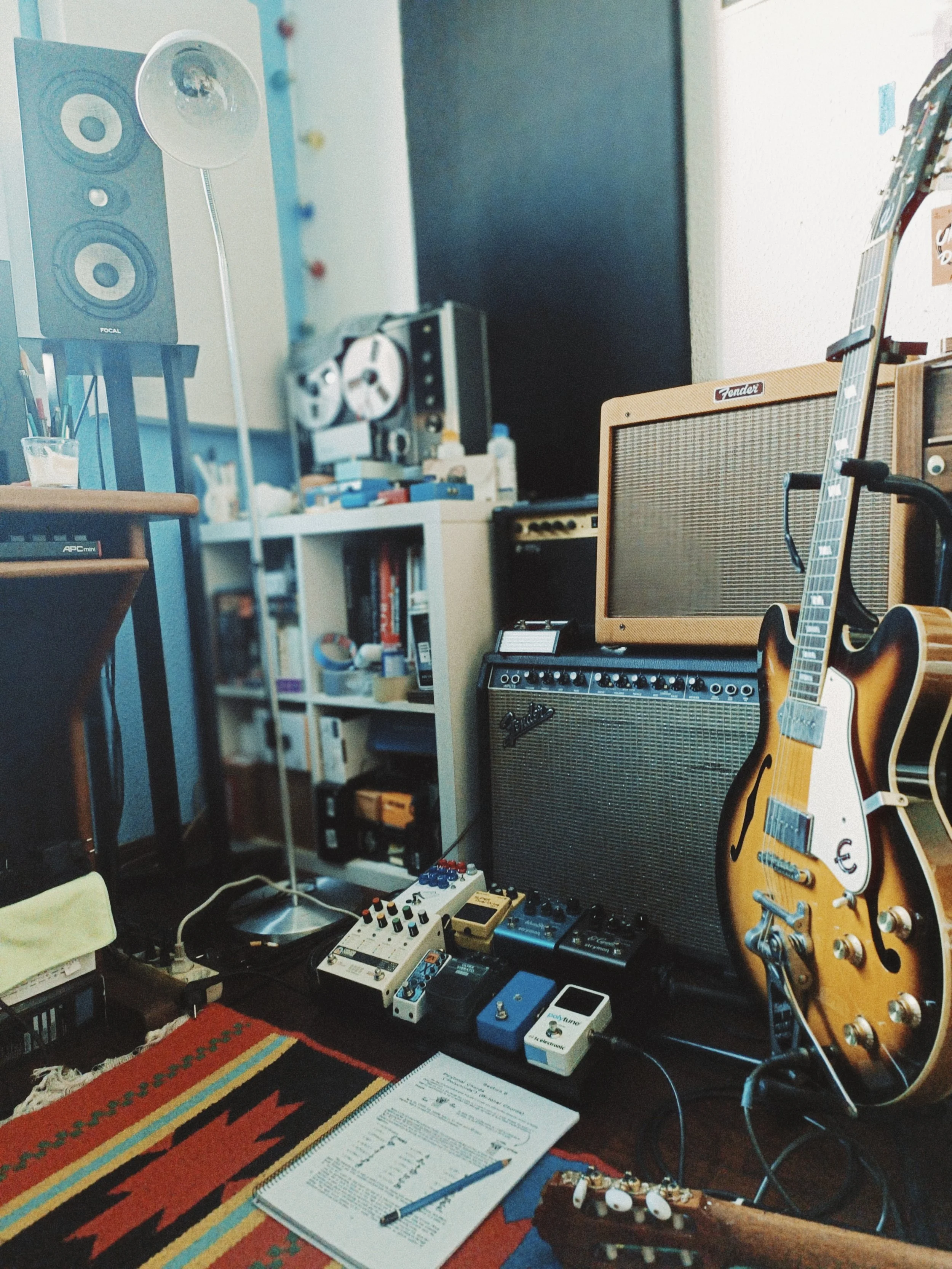 Musical equipment including a guitar, amplifier, multiple effect pedals, a speaker, and a reel-to-reel tape recorder in a room with bookshelves, a lamp, and a colorful rug.