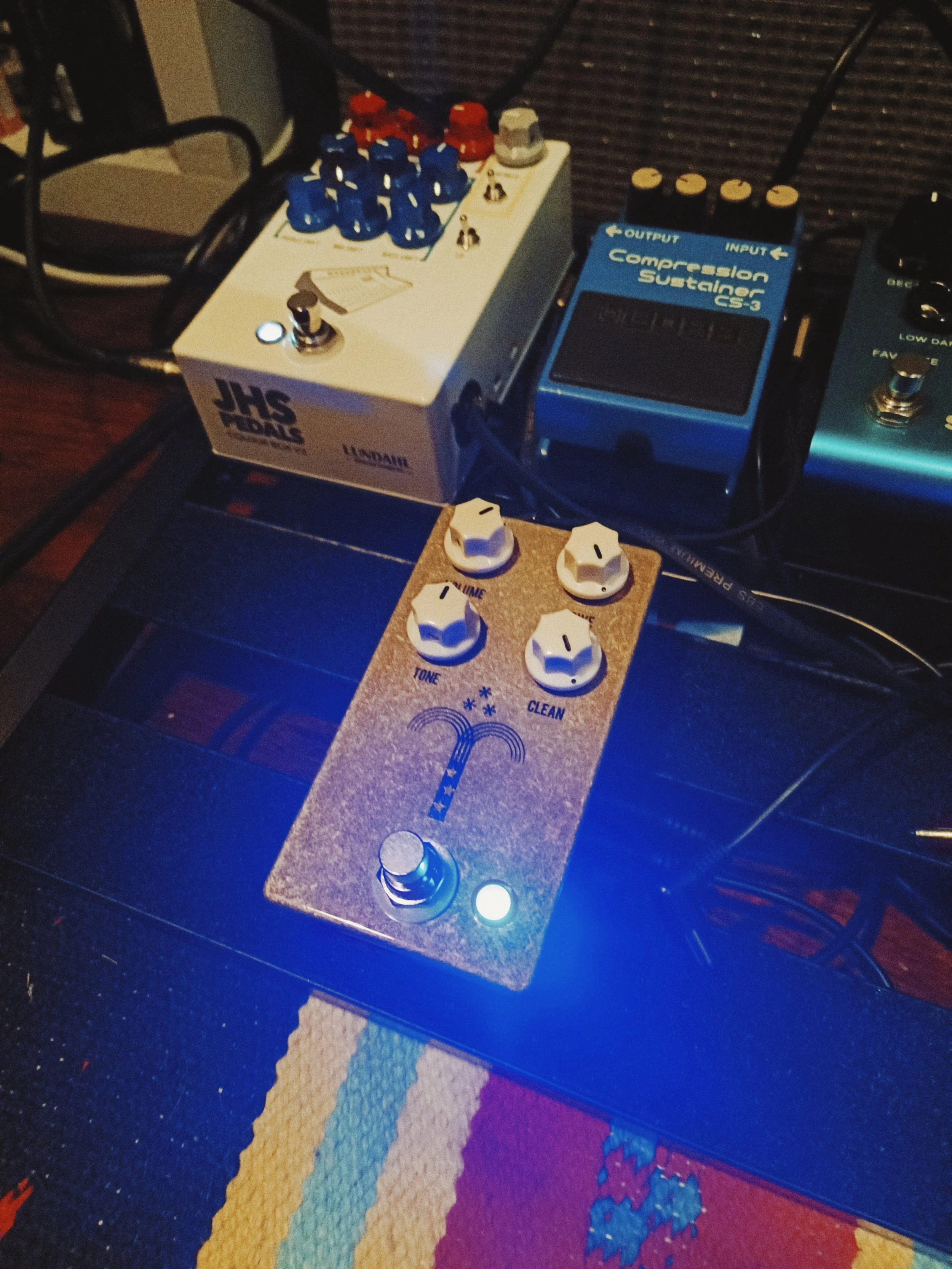 A close-up image of various guitar effects pedals arranged on a pedalboard, including a blue compressor sustain pedal, and a white JHS The Peach pedal with colorful knobs, placed on a multicolored textured surface.