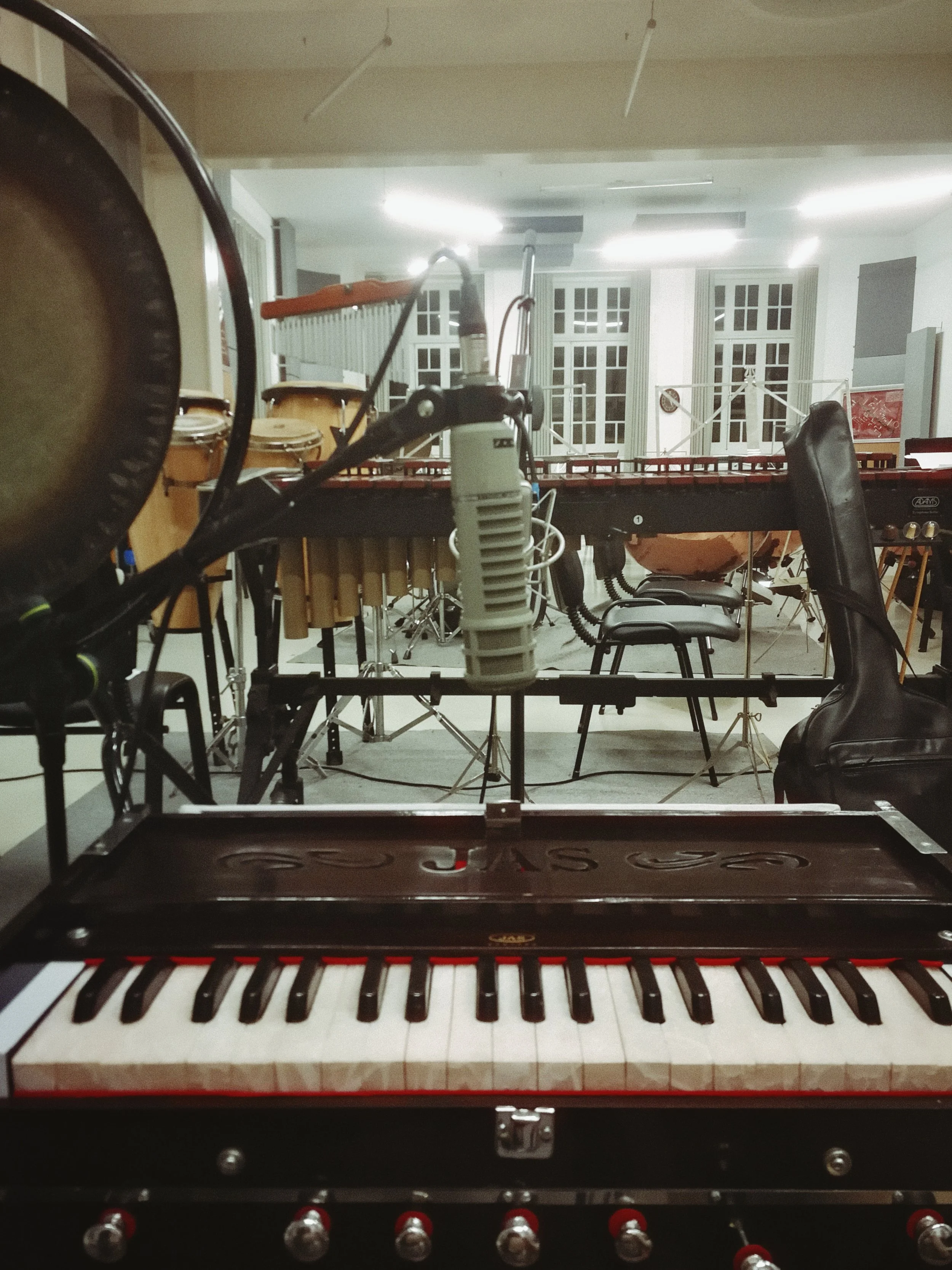 A room with musical instruments including a keyboard in the foreground, drums and percussion instruments in the background, and various chairs and music stands, illuminated by ceiling lights.