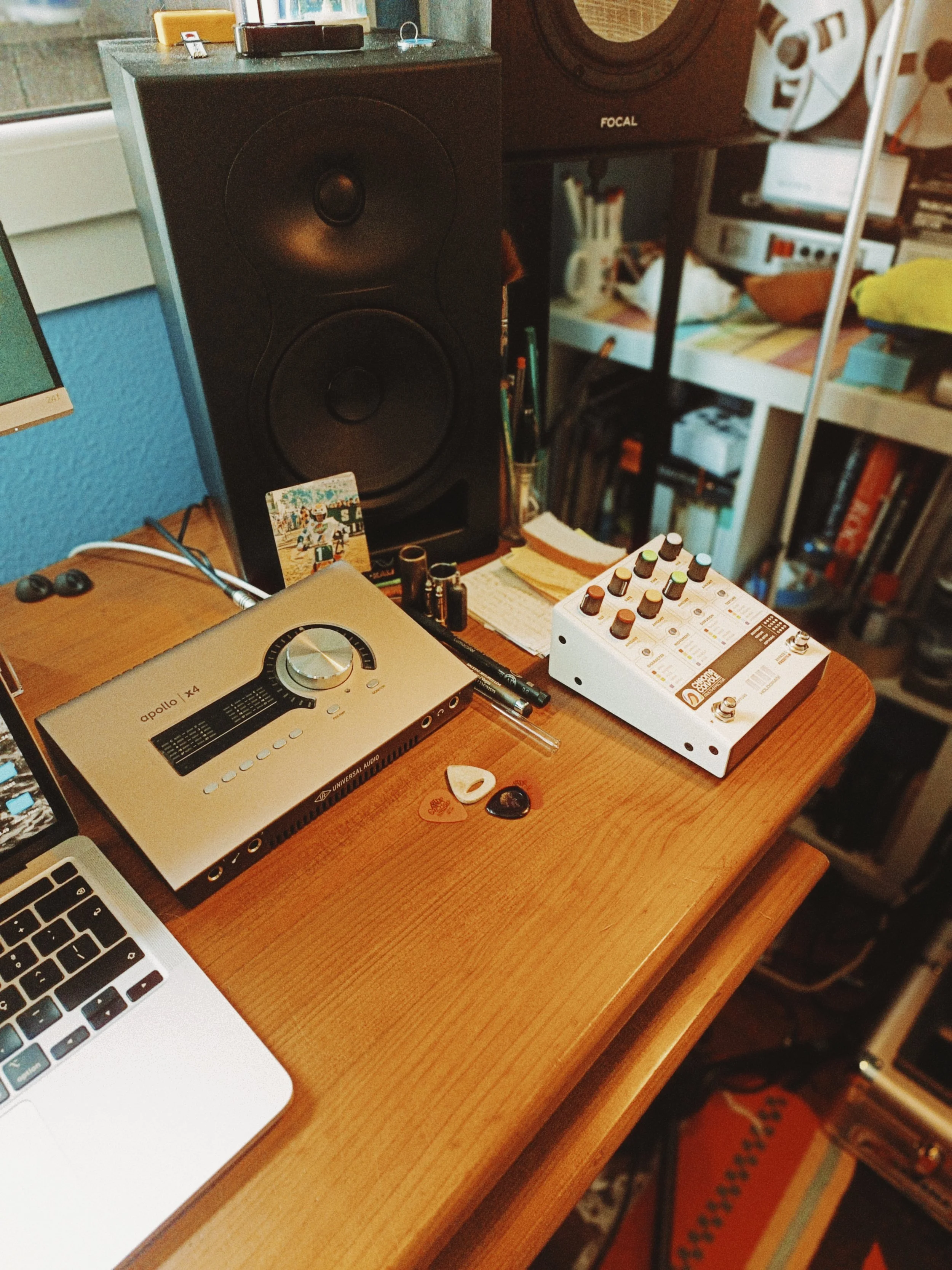 A wooden desk holds a MacBook laptop, a DJ Apollo audio interface, a guitar pick, and some small electronic components. Behind the desk, there are two large speakers, one branded Focal, and a bookcase filled with books and items. A colorful rug is pa