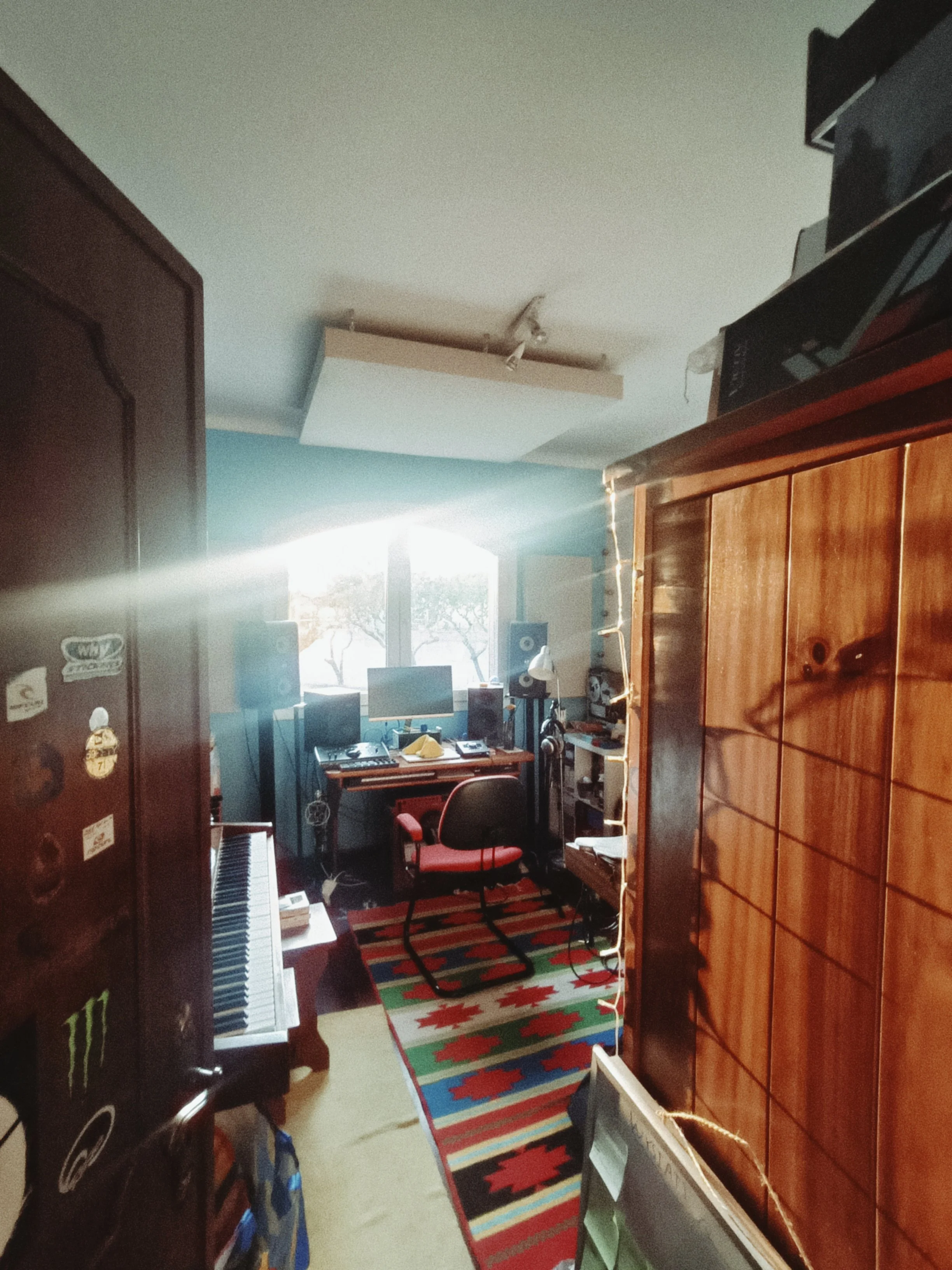 A cluttered room with a desk containing computer monitors, speakers, and a keyboard, a red office chair, a patterned rug, a tall wooden cabinet, and a window with sunlight streaming in.