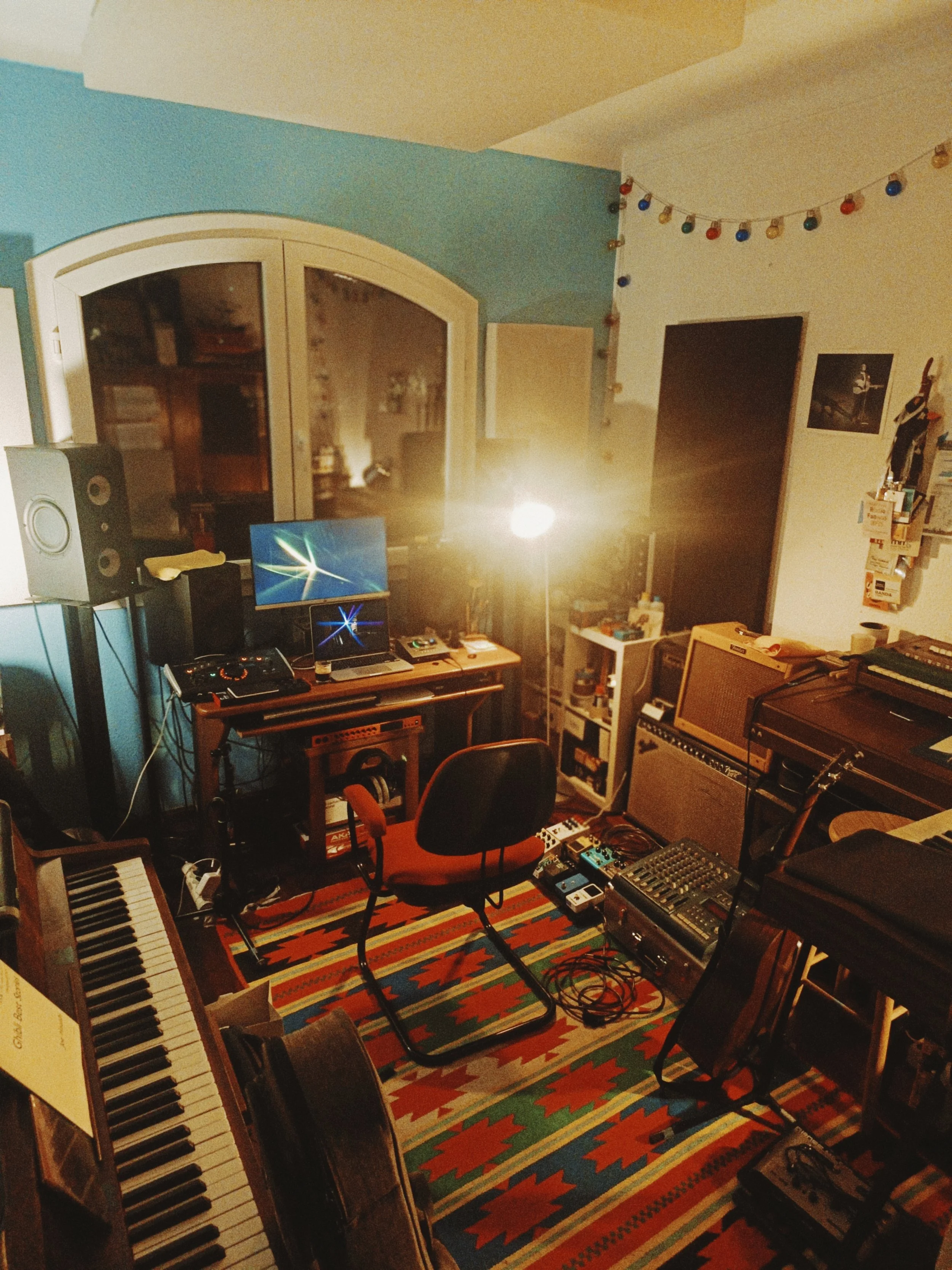 Photo of a music studio or practice room with a keyboard on the left, a colorful geometric patterned rug on the floor, a computer monitor, a chair, and musical equipment, including a drum machine, speakers, and a guitar amplifier, with string lights 