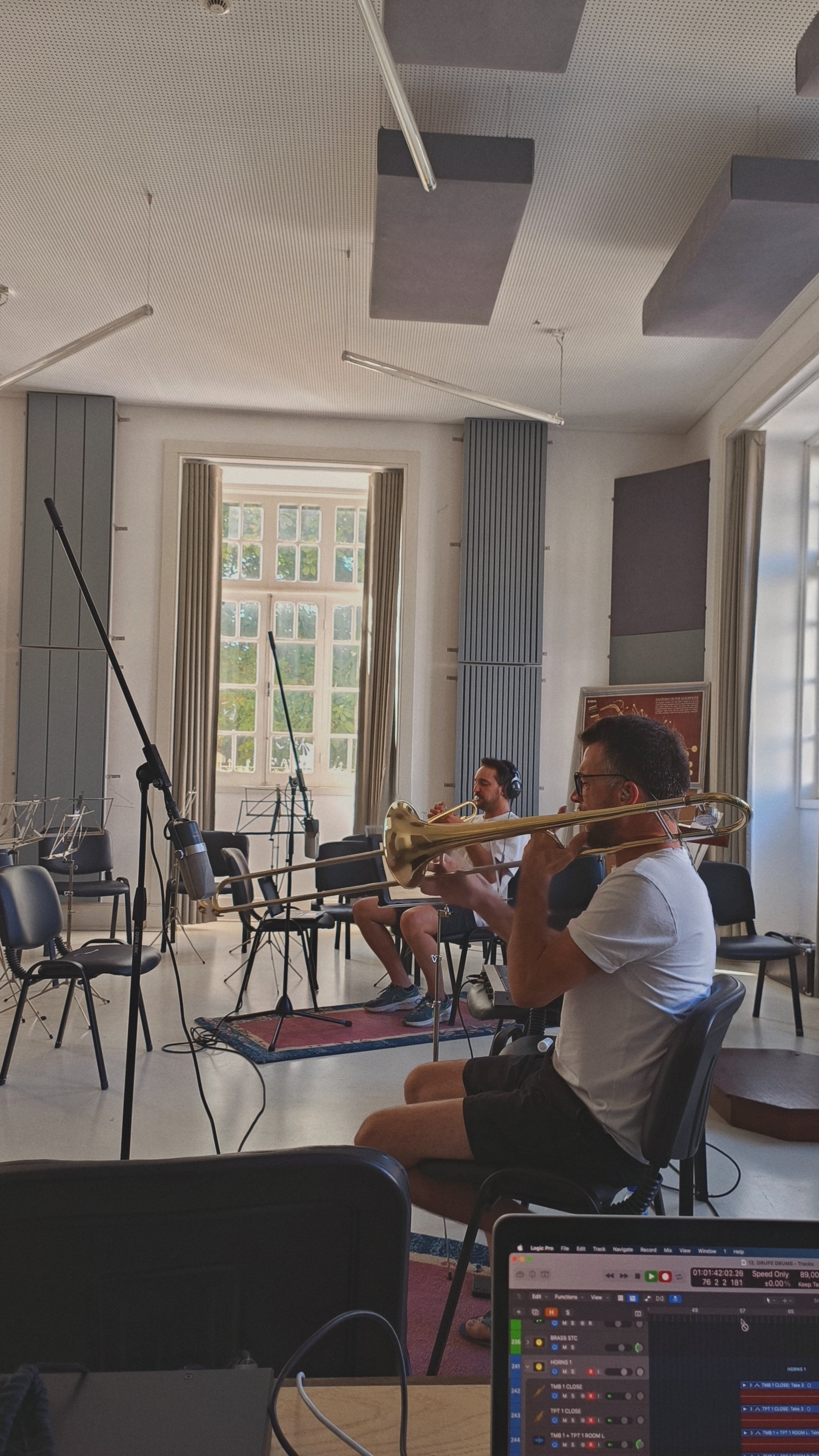 Two musicians seated in a music studio playing brass instruments, with microphones and music stands around them, on a sunny day with large windows.