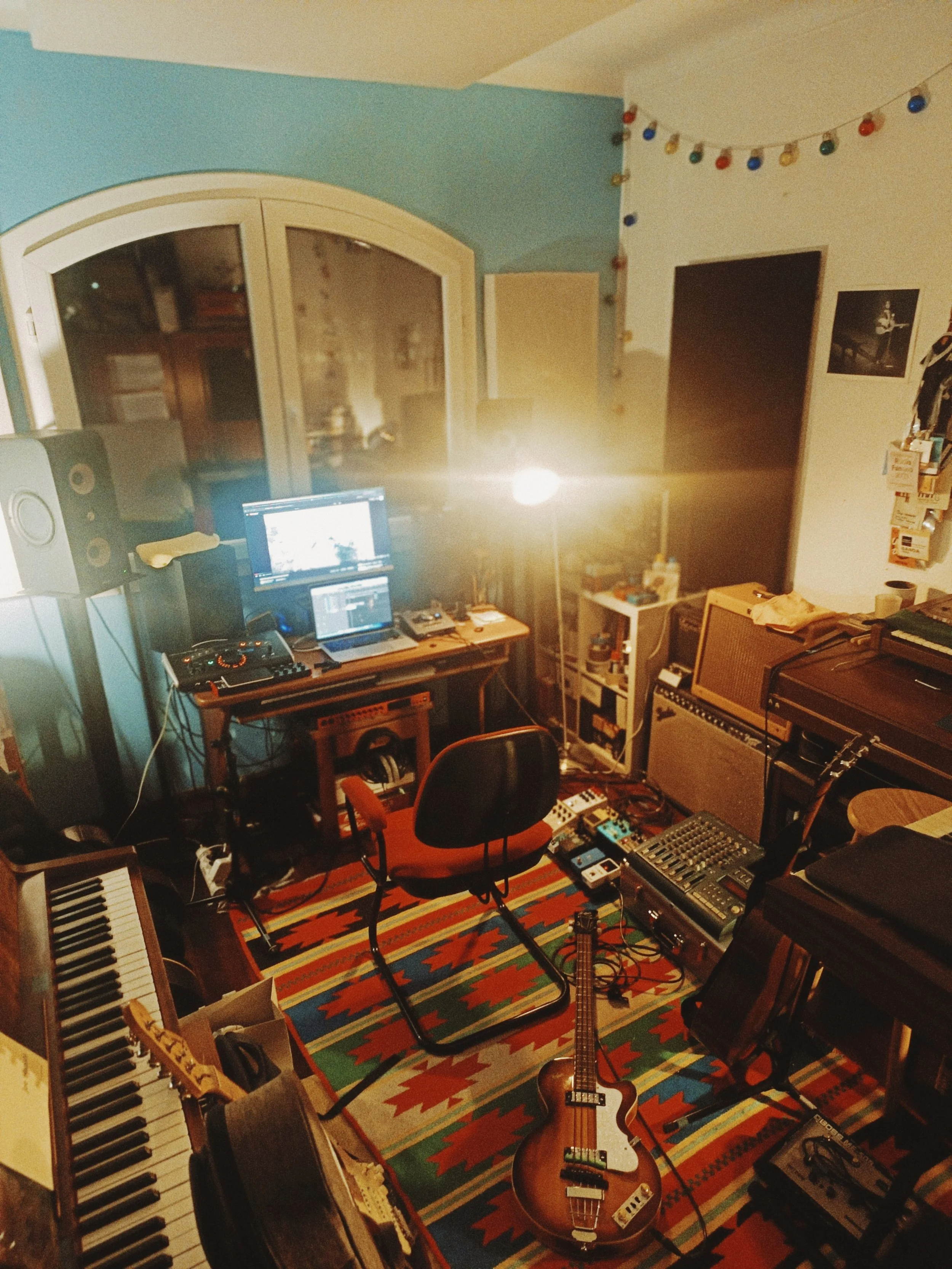 A room with musical instruments, including a keyboard, guitar, and synthesizer, along with a computer, speakers, and various equipment on a colorful patterned rug.