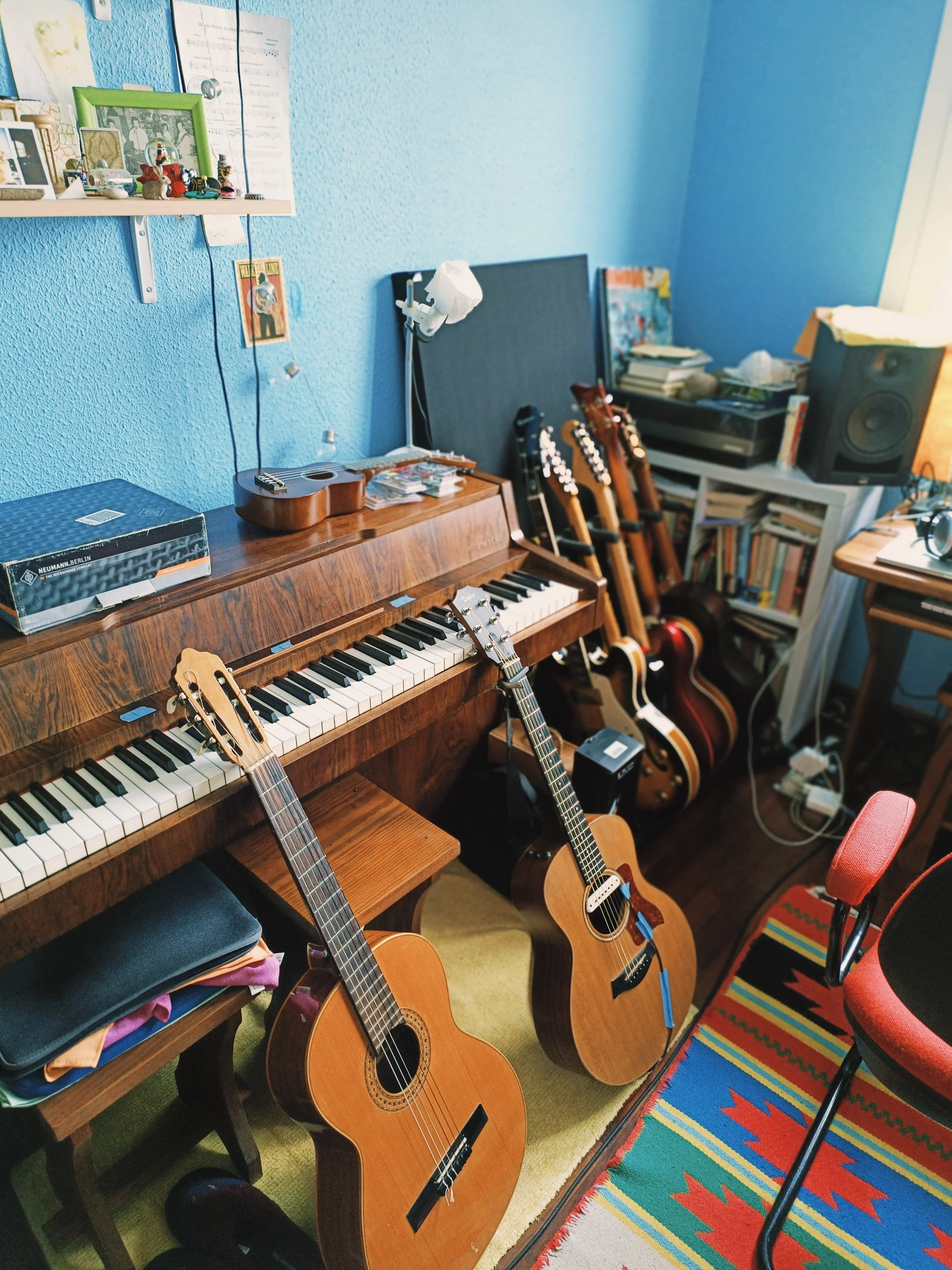 A room with a wooden keyboard, several acoustic guitars, a bookshelf filled with books, a black speaker, a small table, and a colorful rug. The blue wall has various pictures and notes pinned or hanging on it.