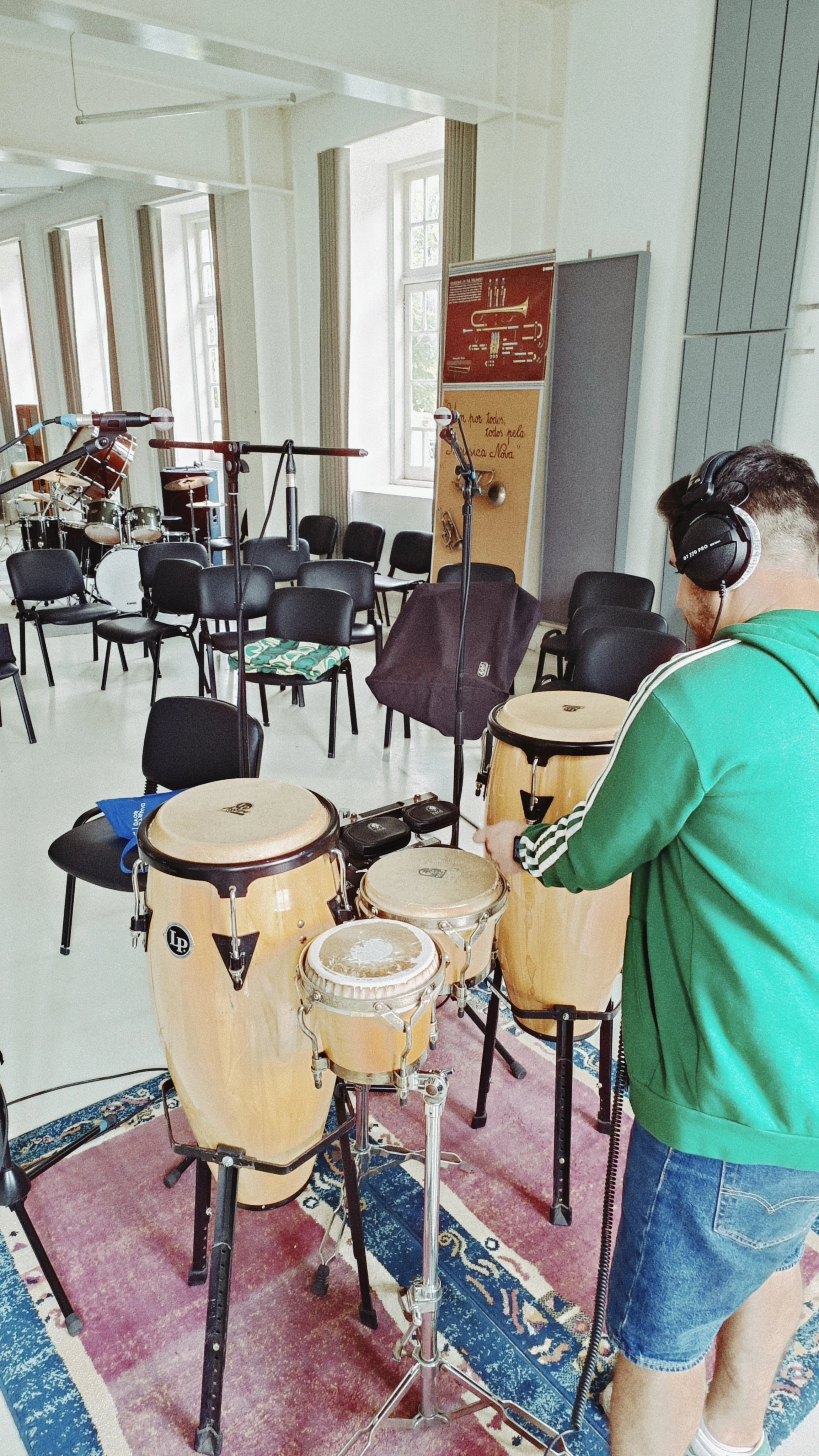 A man in a green jacket and denim shorts is playing conga drums in a room with chairs, microphones, and musical instruments, preparing for a performance.