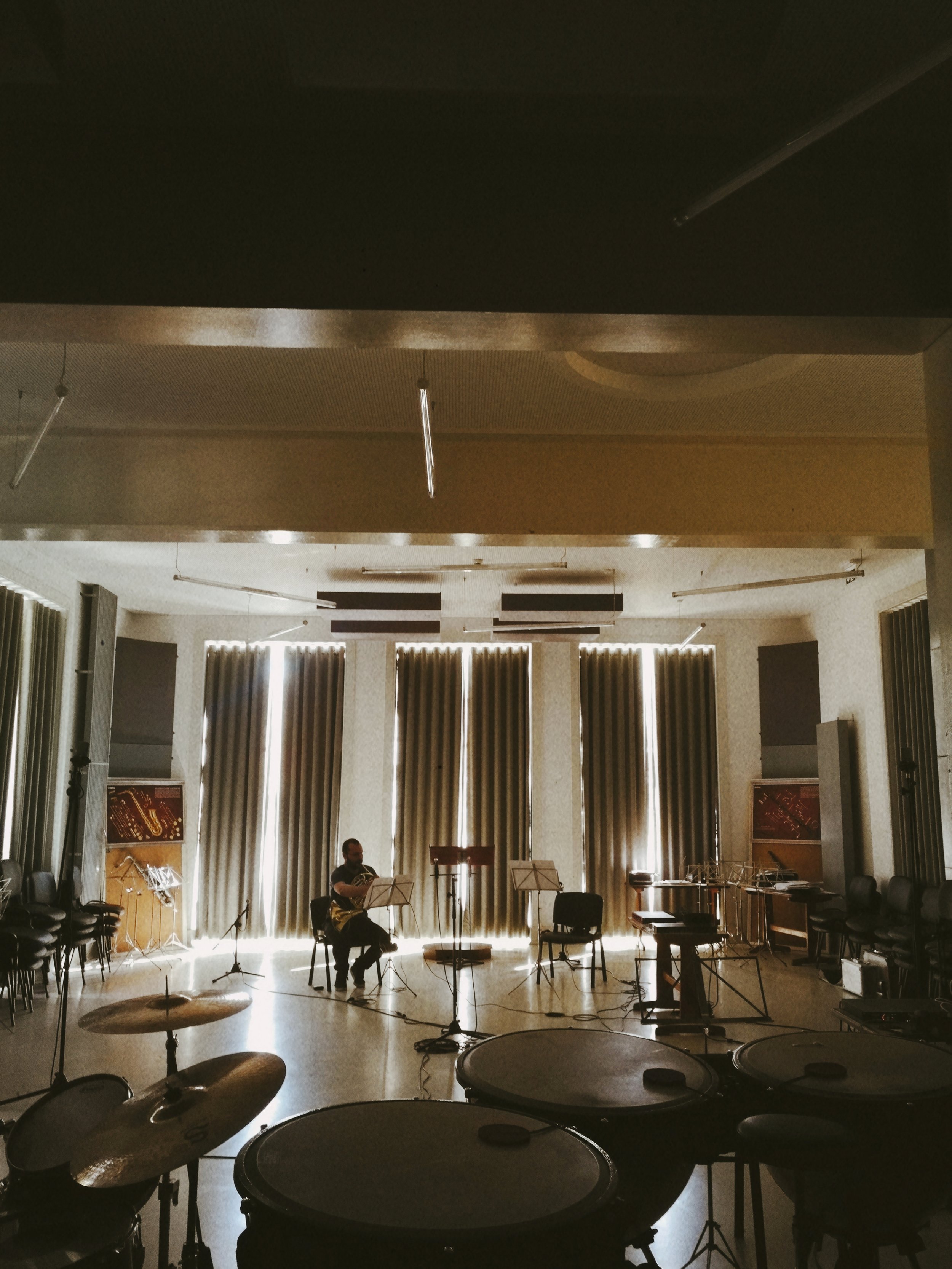 An indoor music rehearsal space with curtains, chairs, and a person playing guitar on stage. Musical instruments and equipment are scattered across the room.