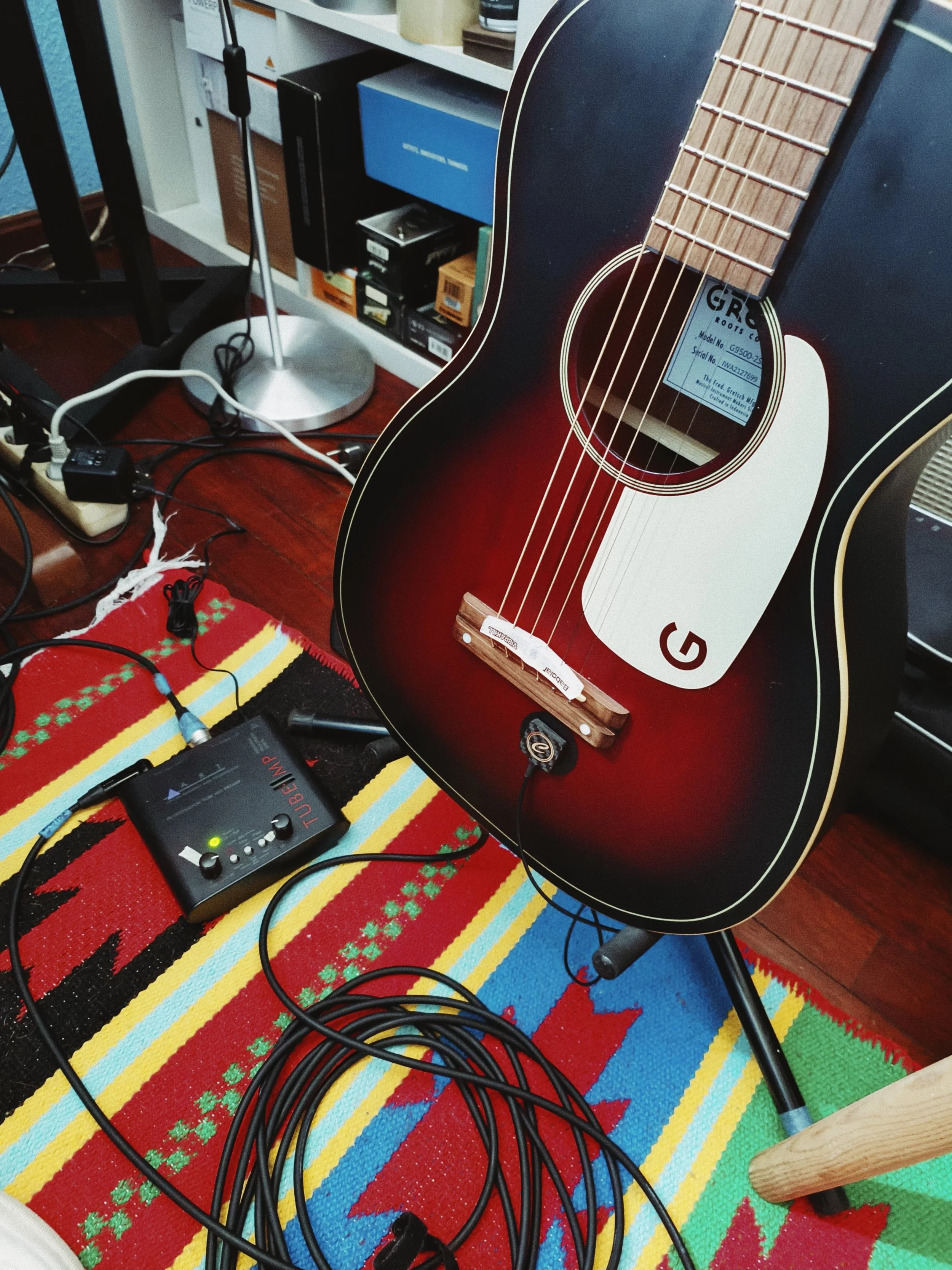 Close-up of a guitar with a dark red sunburst finish on a colorful striped rug, connected to an electronic device with multiple black cables.