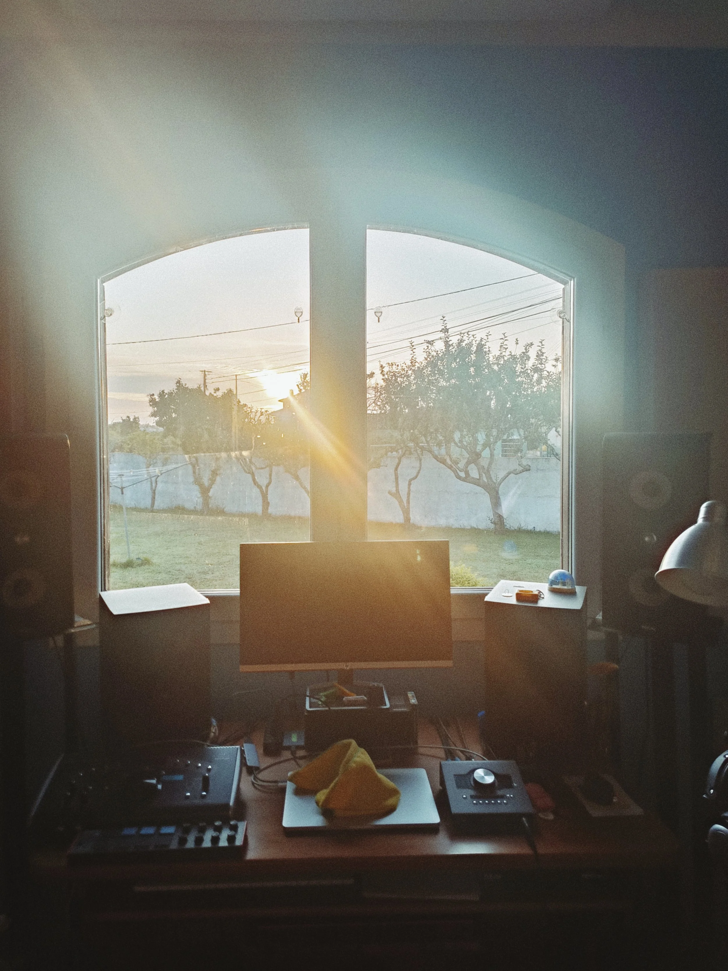 A view of a computer setup on a desk with DJ equipment, including a mixer and a turntable, in front of a window showing outdoor trees and power lines during sunset.