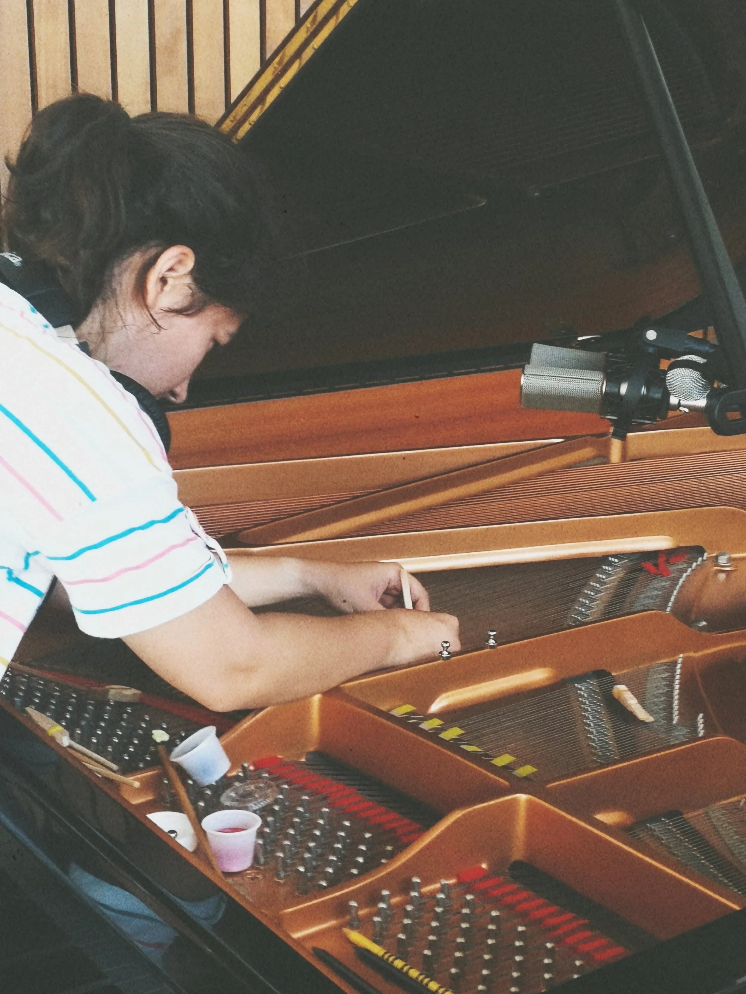 A person with dark hair in a bun wearing a striped shirt leaning over a grand piano with its lid open, revealing the strings and hammers inside, in a wood-paneled room.