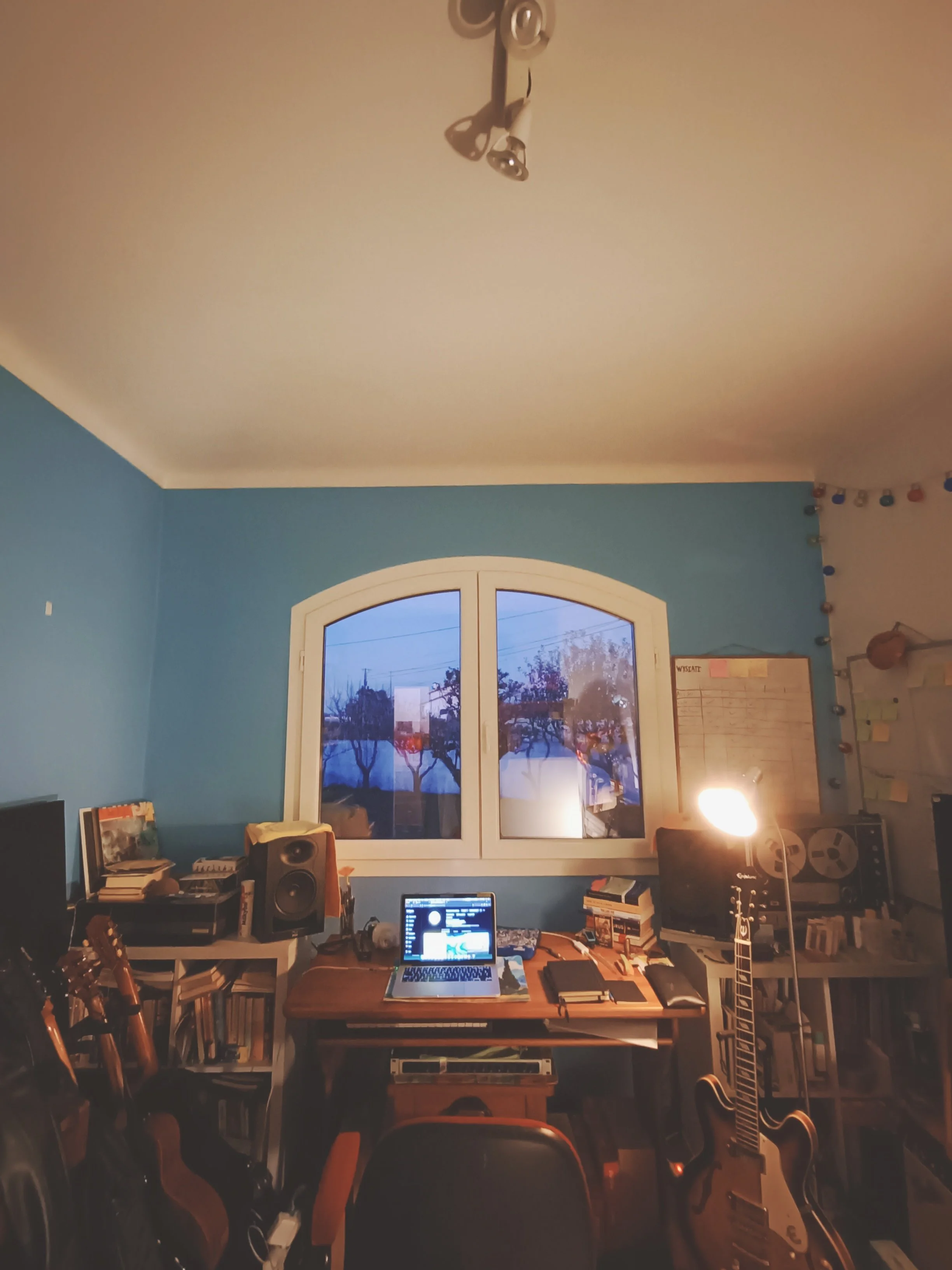 A cozy home office with a wooden desk, a computer, speakers, and musical instruments including a guitar and a ukulele. There is a window with a tree view, bookshelves filled with books, and string lights along the wall. The room is warmly lit with a 