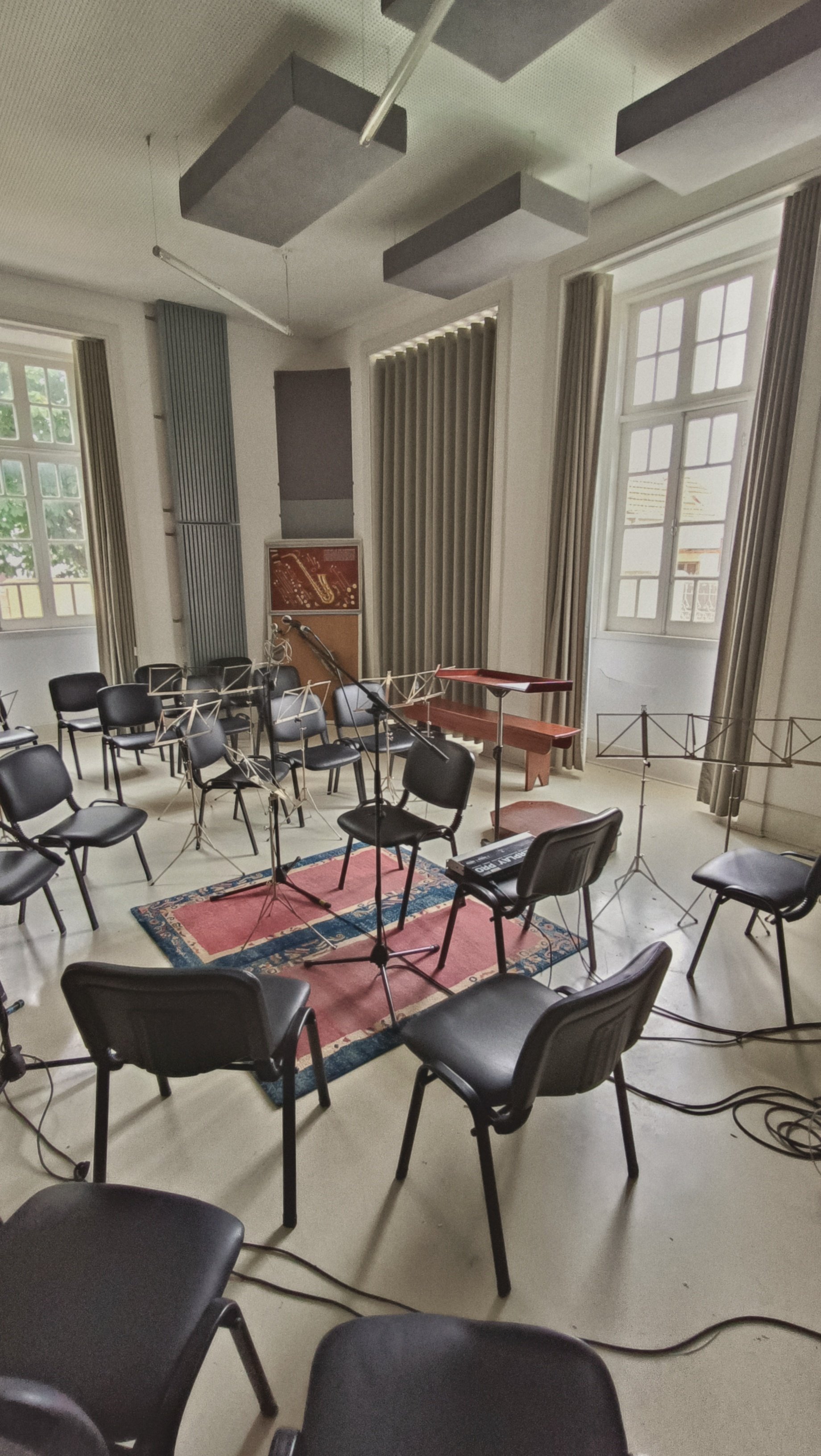 Empty music rehearsal room with chairs, music stands, a piano, and a microphone, set up for a band or orchestra, with large windows and ceiling sound panels.