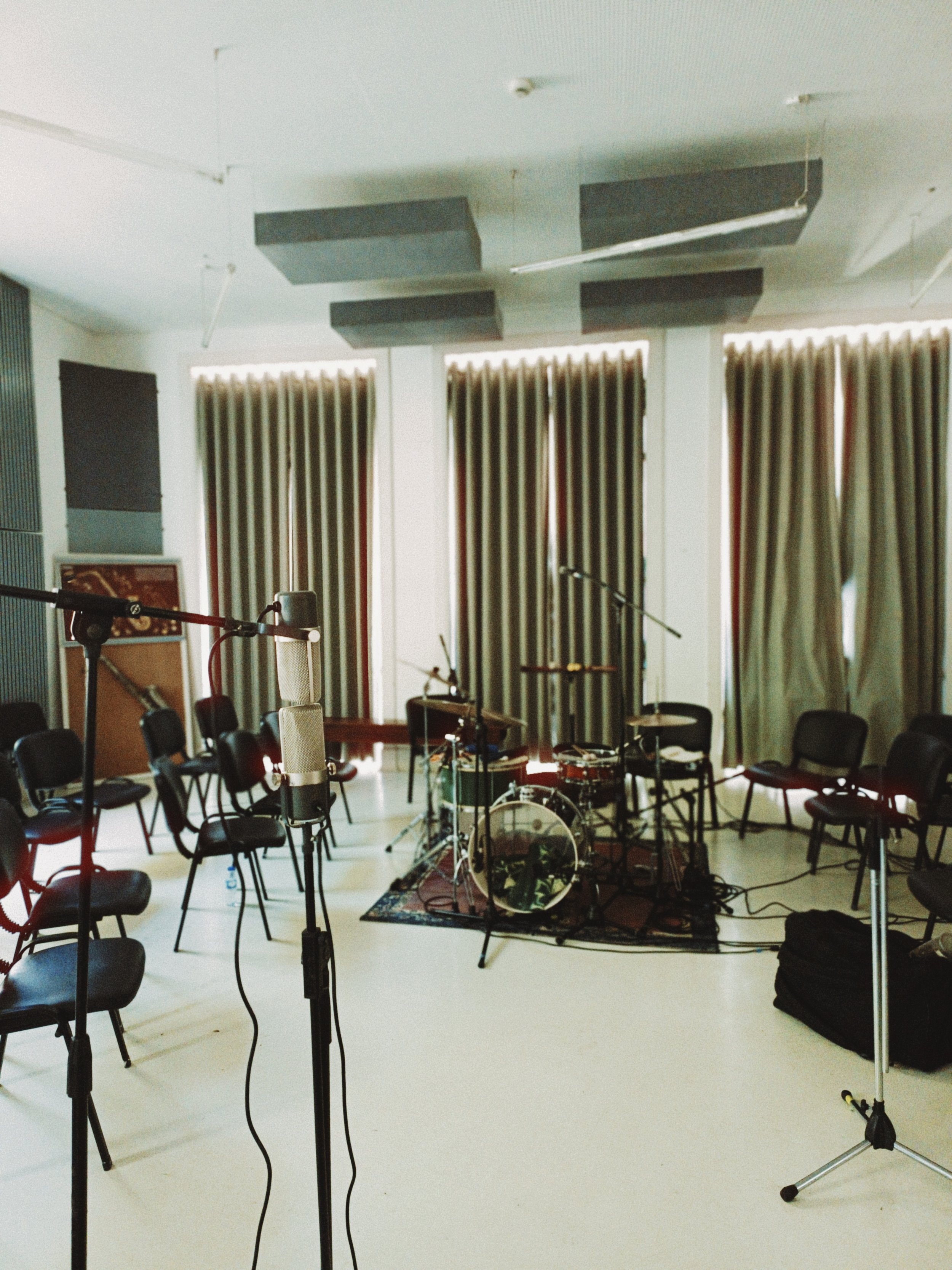 A music rehearsal or recording studio with microphones, chairs, and a drum set arranged on a rug, with large windows and curtains in the background.