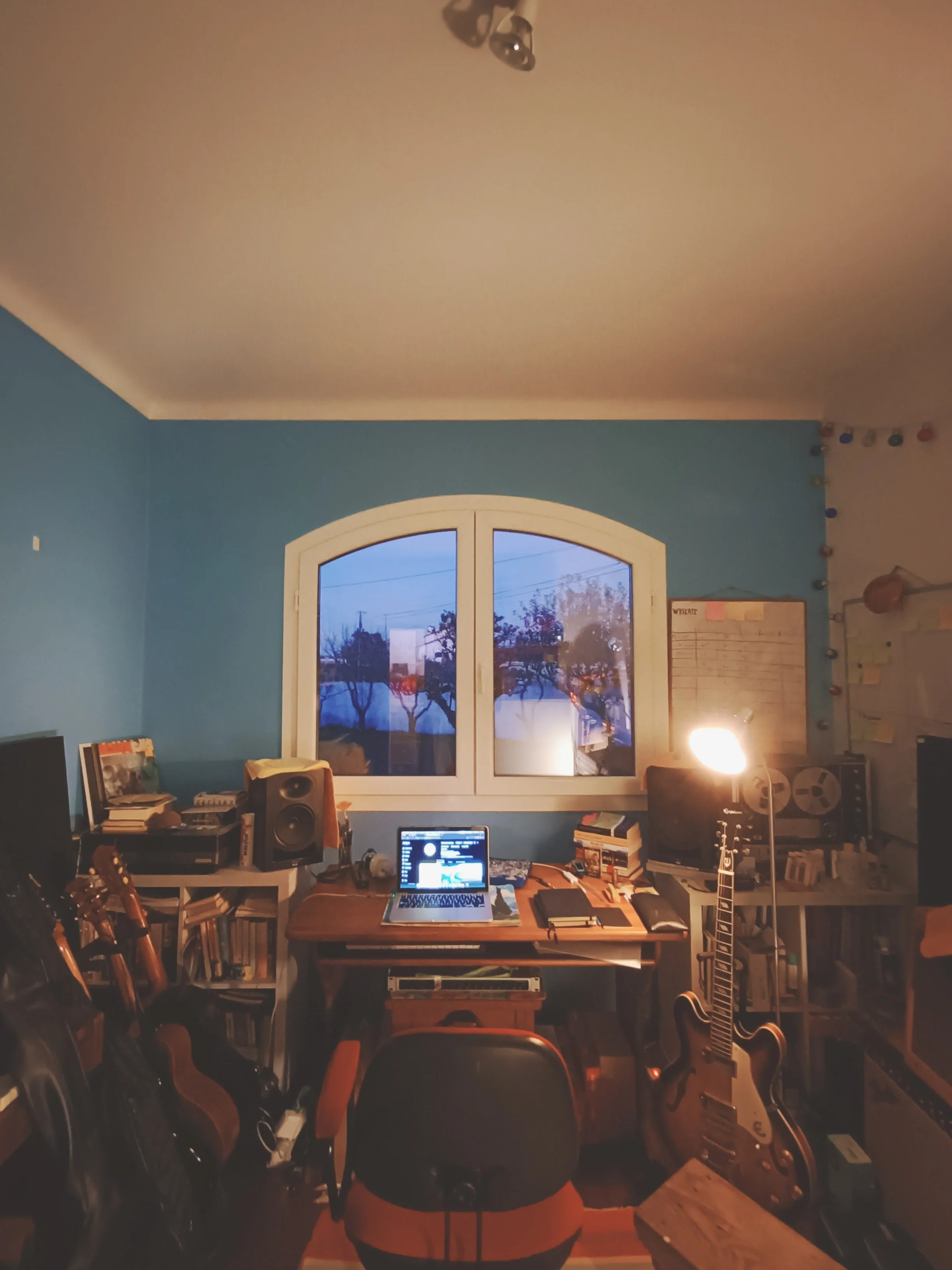 A home music studio with a desk filled with a laptop, speakers, and musical equipment, positioned in front of a large window showing trees outside. The room has blue walls, bookshelves, and studio gear, with guitars on stands and a projector on a sid