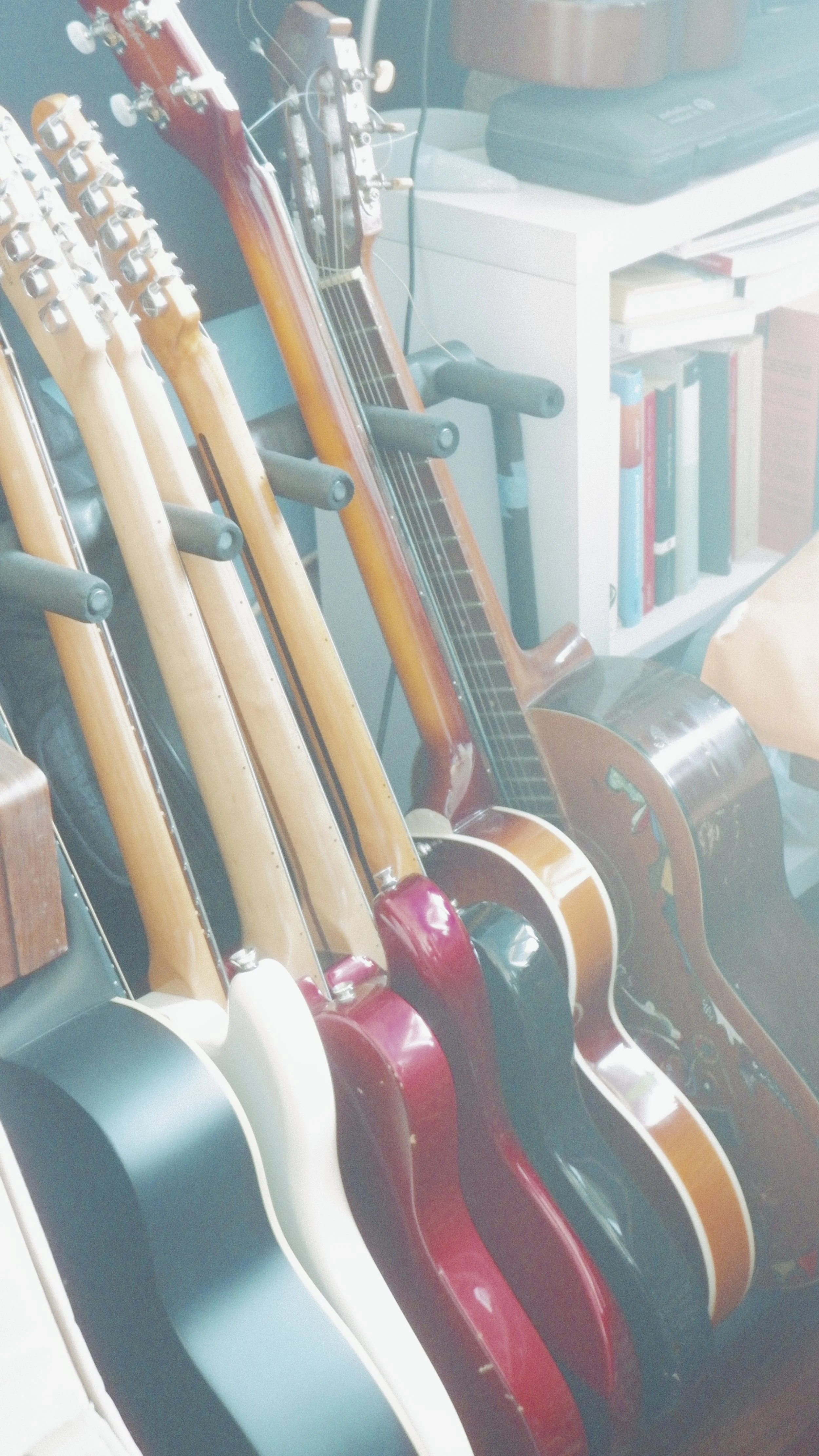 A collection of various colored guitars and a banjo organized on a rack near a white bookshelf with books and a laptop on top.