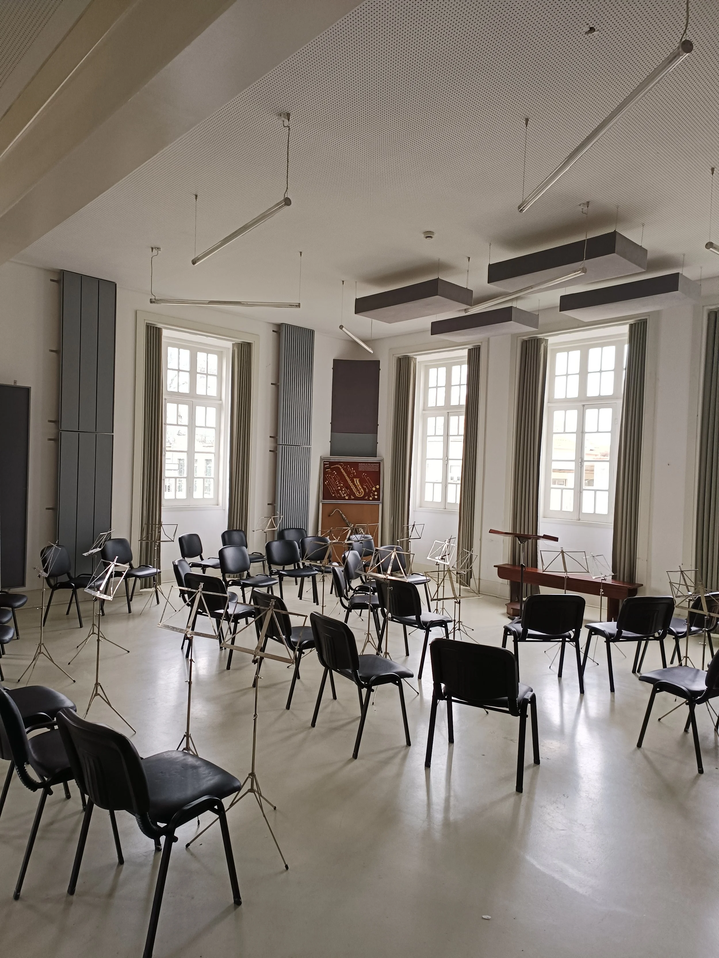 An empty classroom or music room with black chairs arranged in a semi-circle, music stands, large windows with curtains, a piano, and soundproofing panels on the walls.