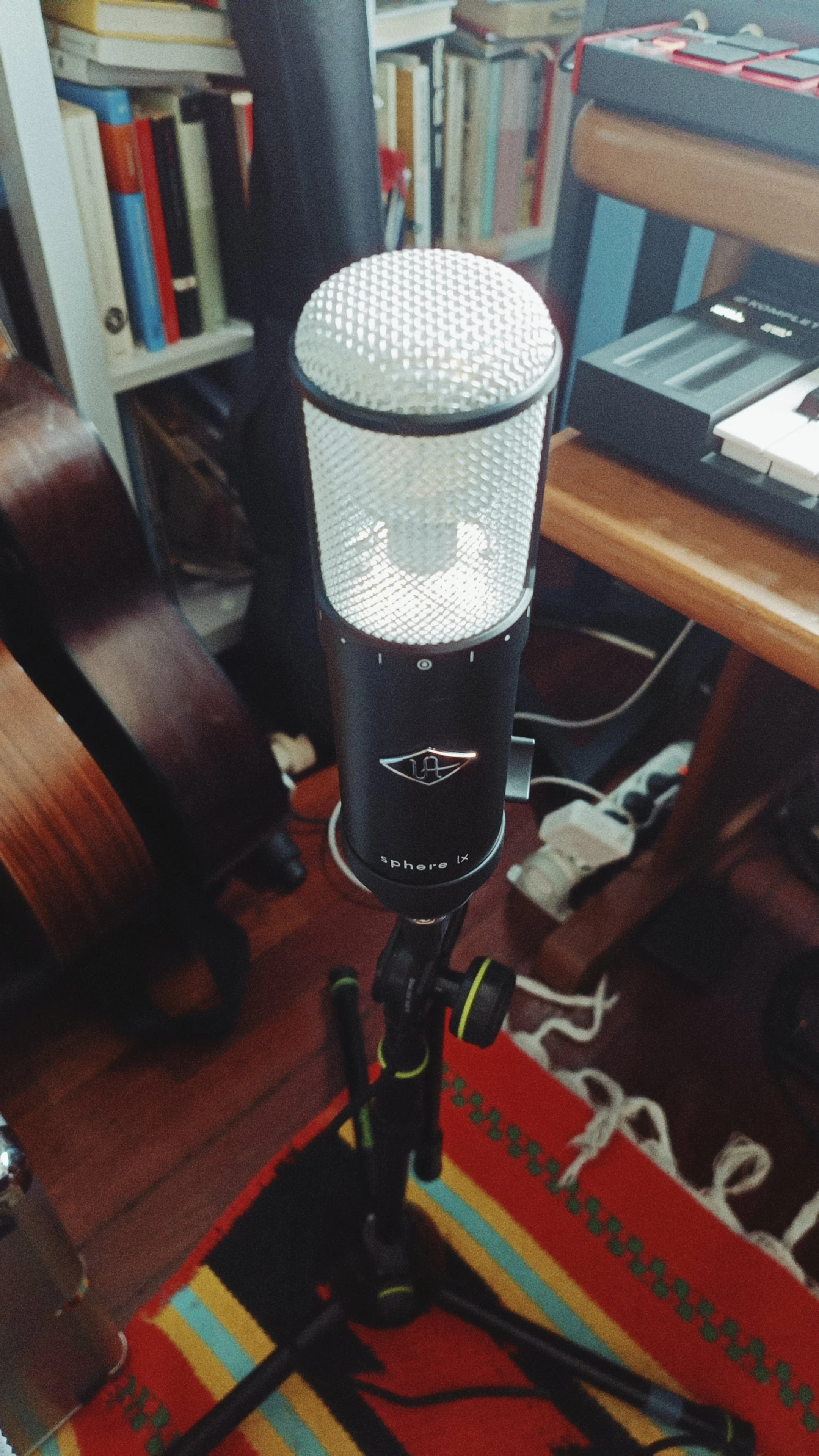 A microphone on a black stand with a textured silver grille, positioned in a room with bookshelves, a keyboard, and a colorful rug.