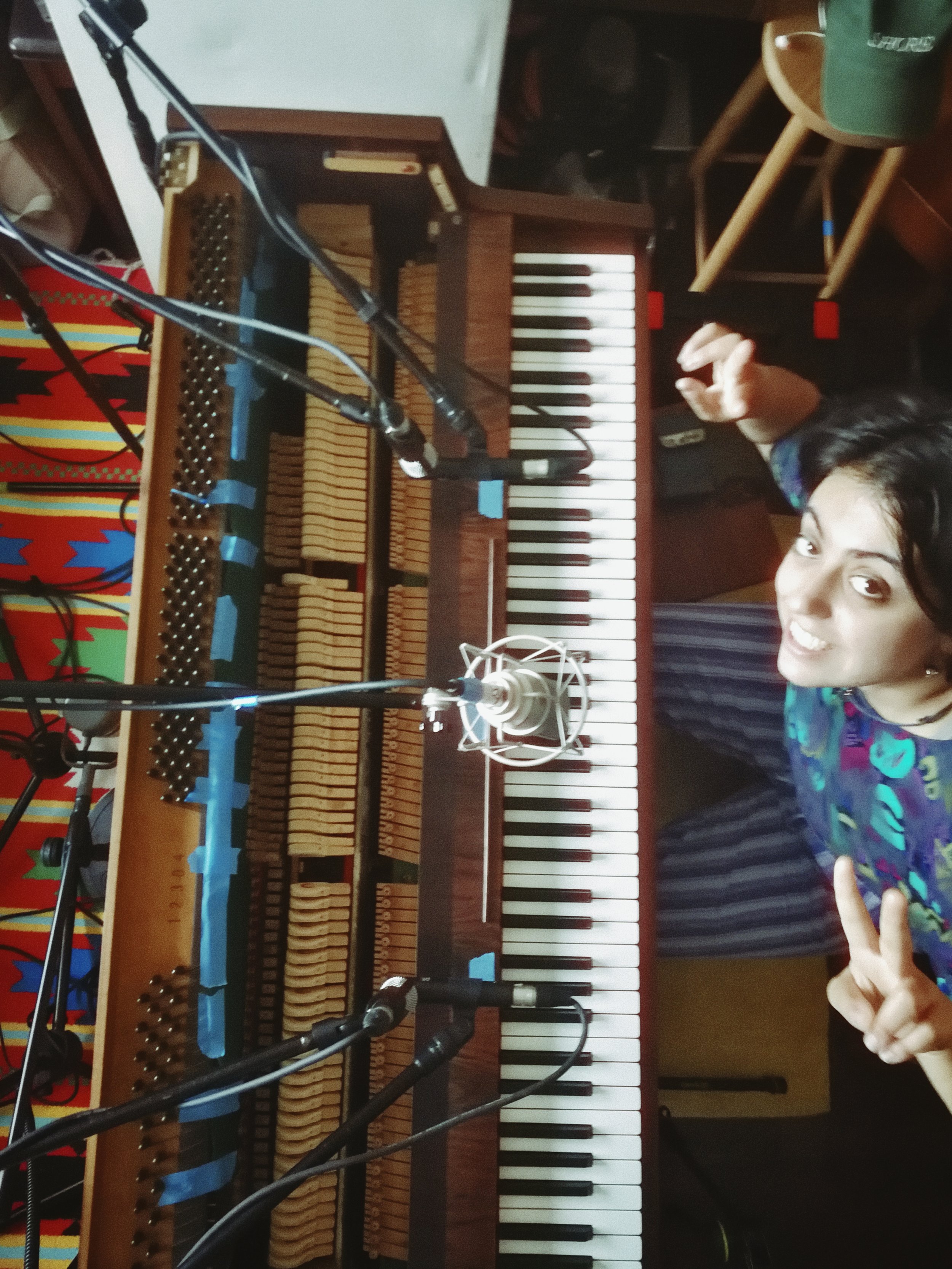 A person with dark hair smiling and making an 'OK' gesture with their right hand, standing in front of an upright piano with a partially opened lid, around microphones.