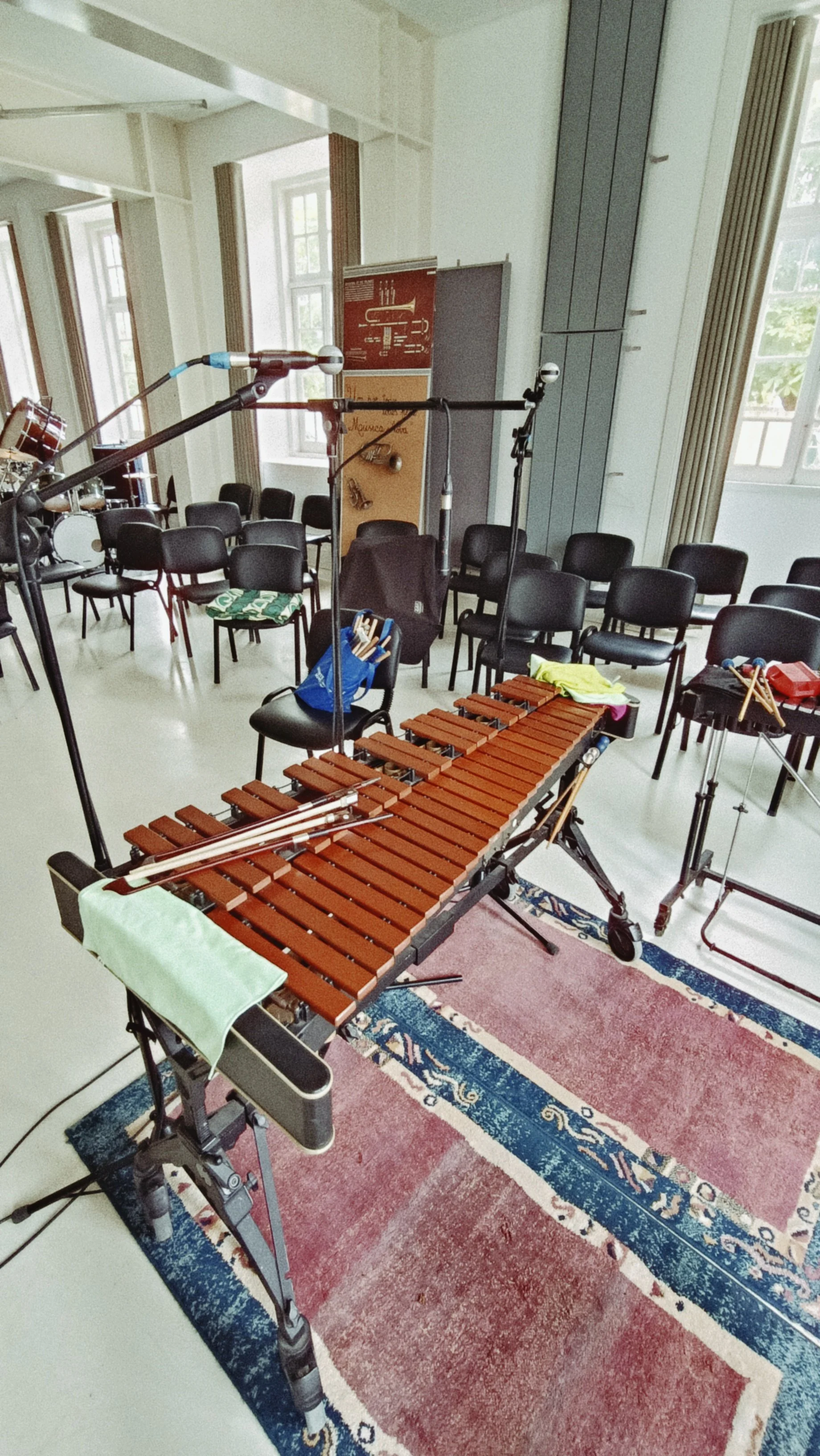 Room set up for a musical performance with a marimba, microphones, and chairs, with windows and curtains in the background.