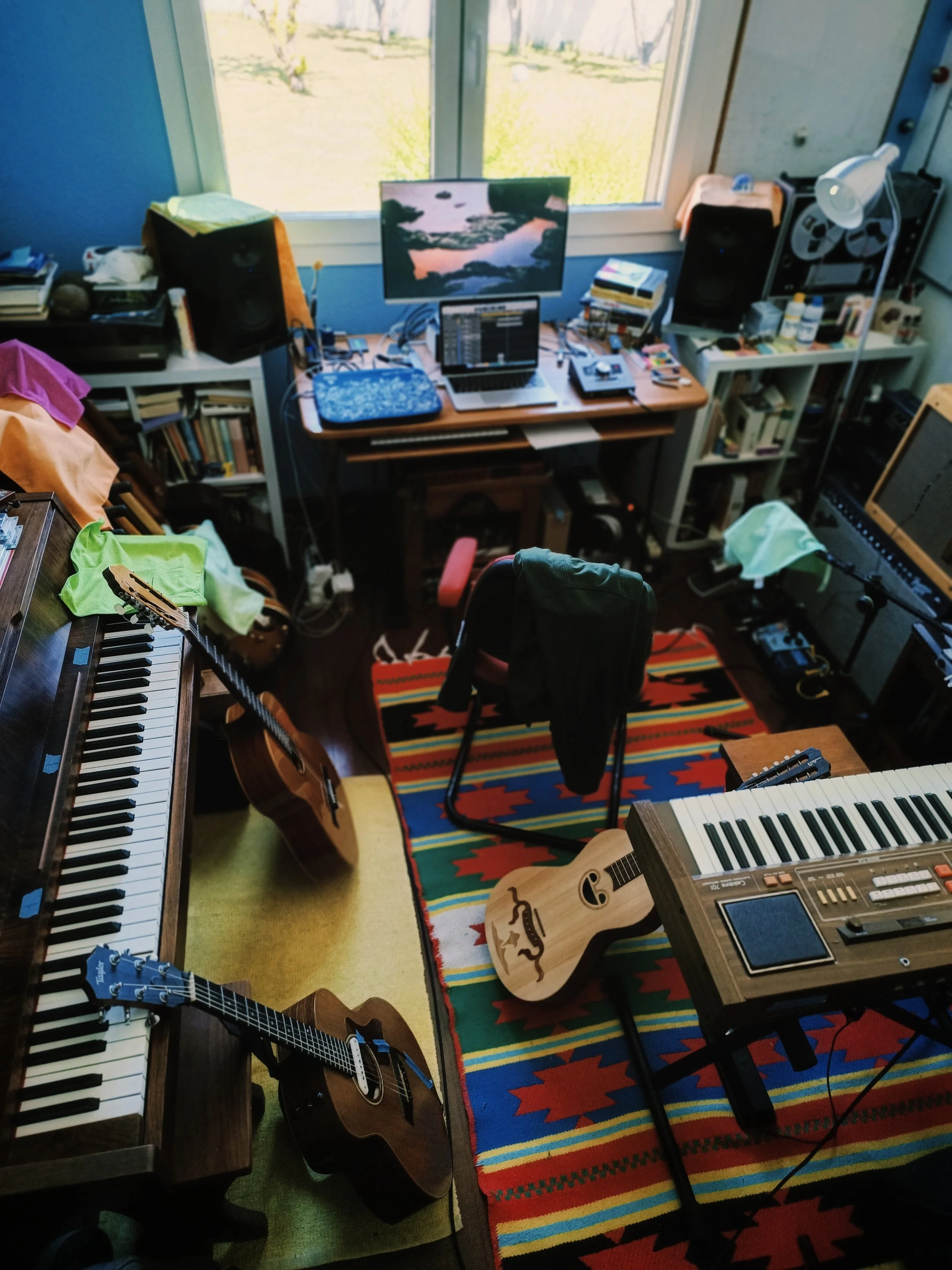 A cluttered music studio with a wooden acoustic guitar on a stand, a portable keyboard, a laptop with audio equipment, a desktop monitor, speakers, books, and various musical accessories on shelves and tables near a window showing a green yard outsid